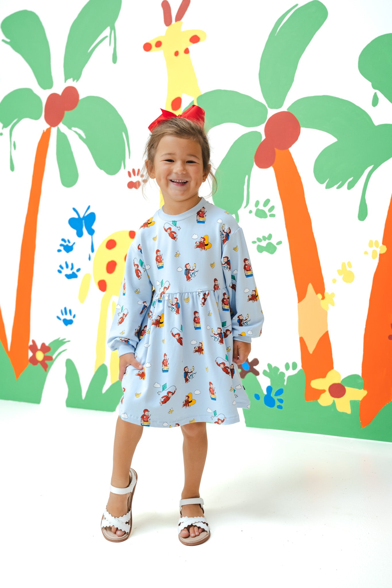 Girl posing in Long Sleeve Skater Dress in Curious George print with a red bow on head standing in front of a colorful background