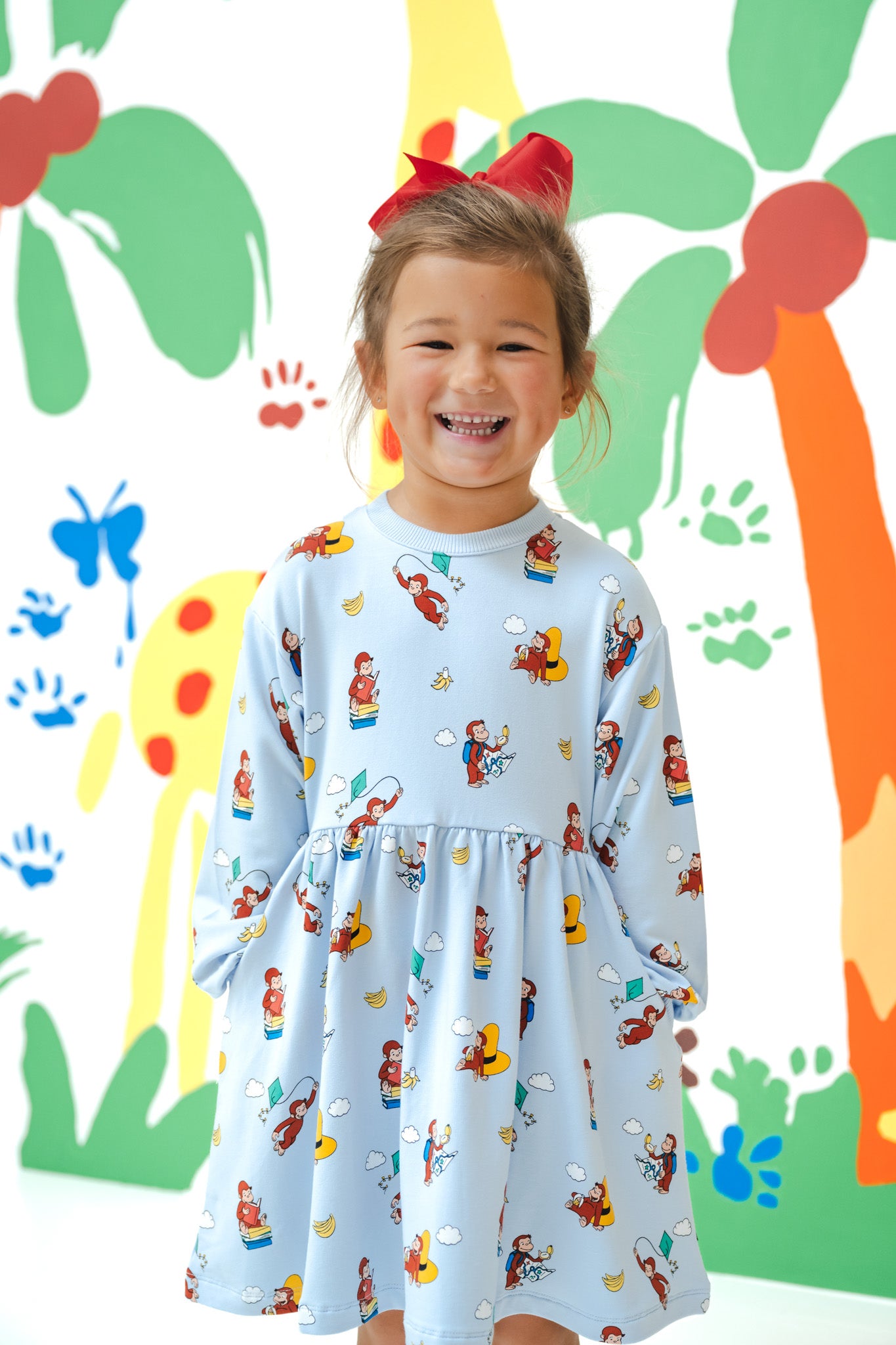 Girl smiling in Long Sleeve Skater Dress in Curious George print with a red bow on head standing in front of a colorful background