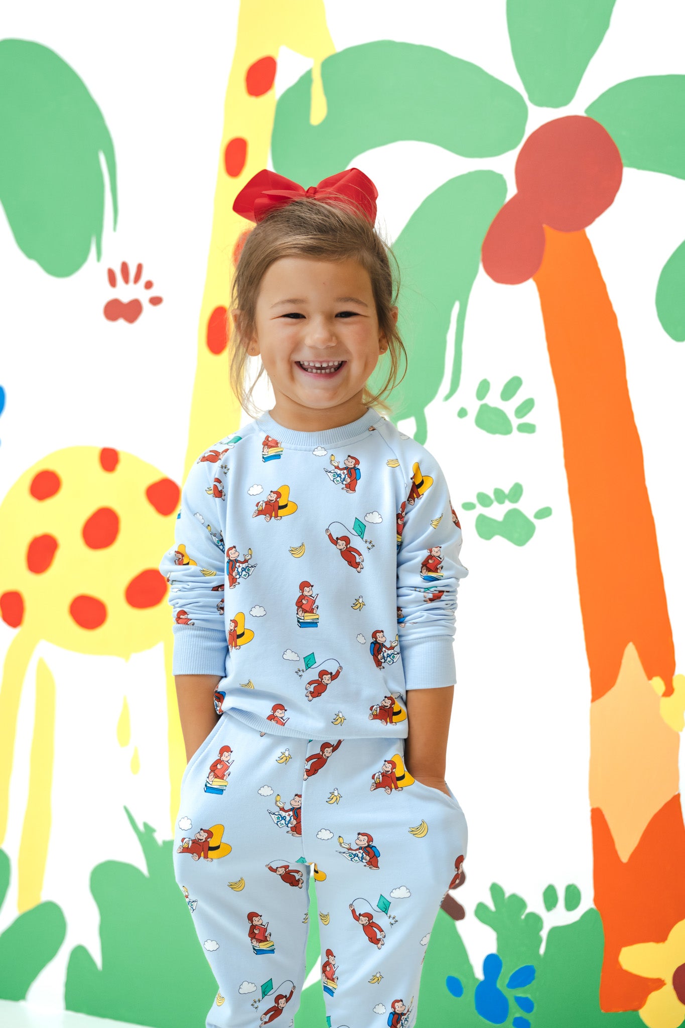 Girl smiling in Long Sleeve Jogger Set in Curious George print standing in front of a colorful background