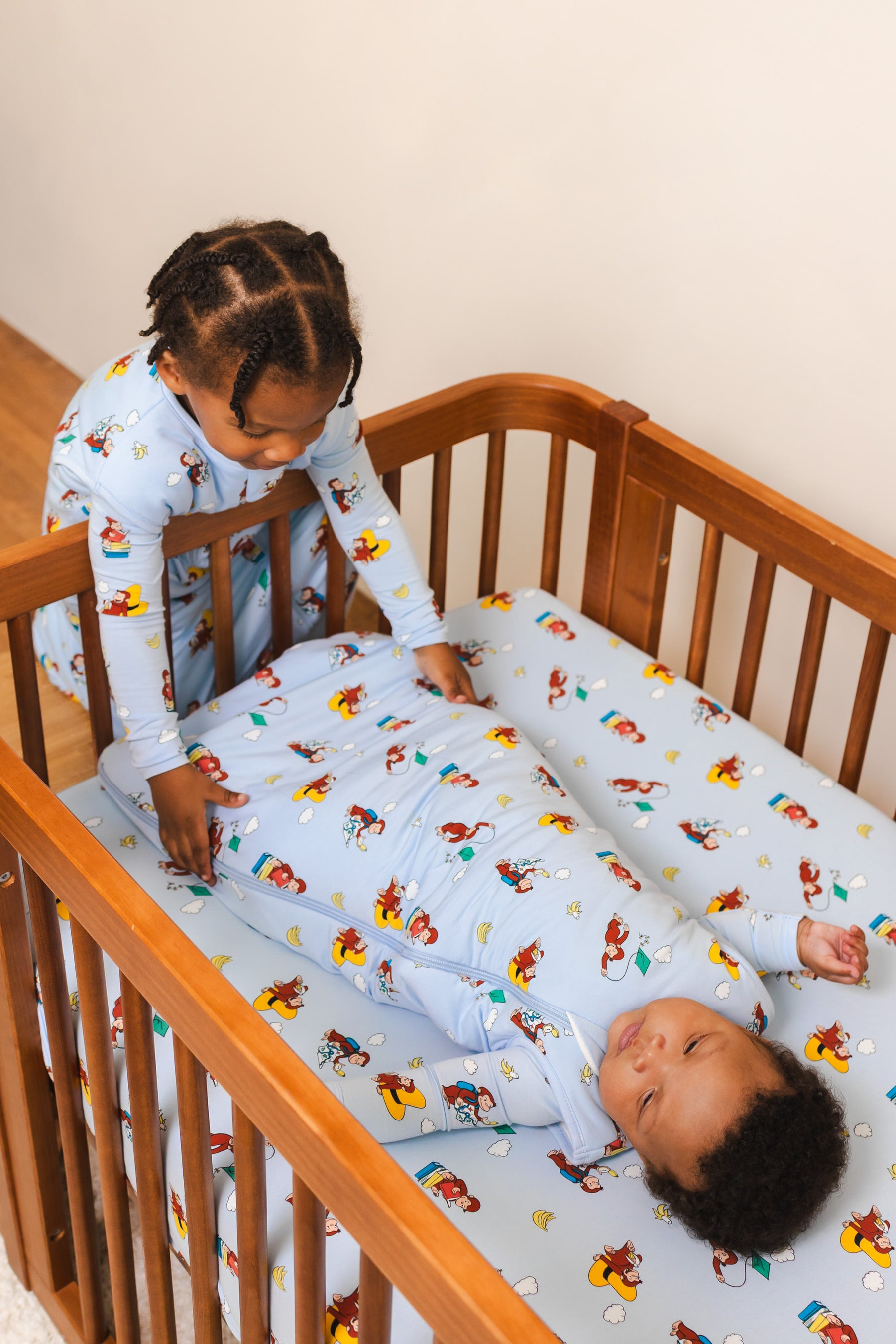Baby lying in Crib in Sleep Bag in Curious George 1.0 print lying on a similar print crib sheet and a girl sitting outside the crib wearing Curious George print Sleep bag walker