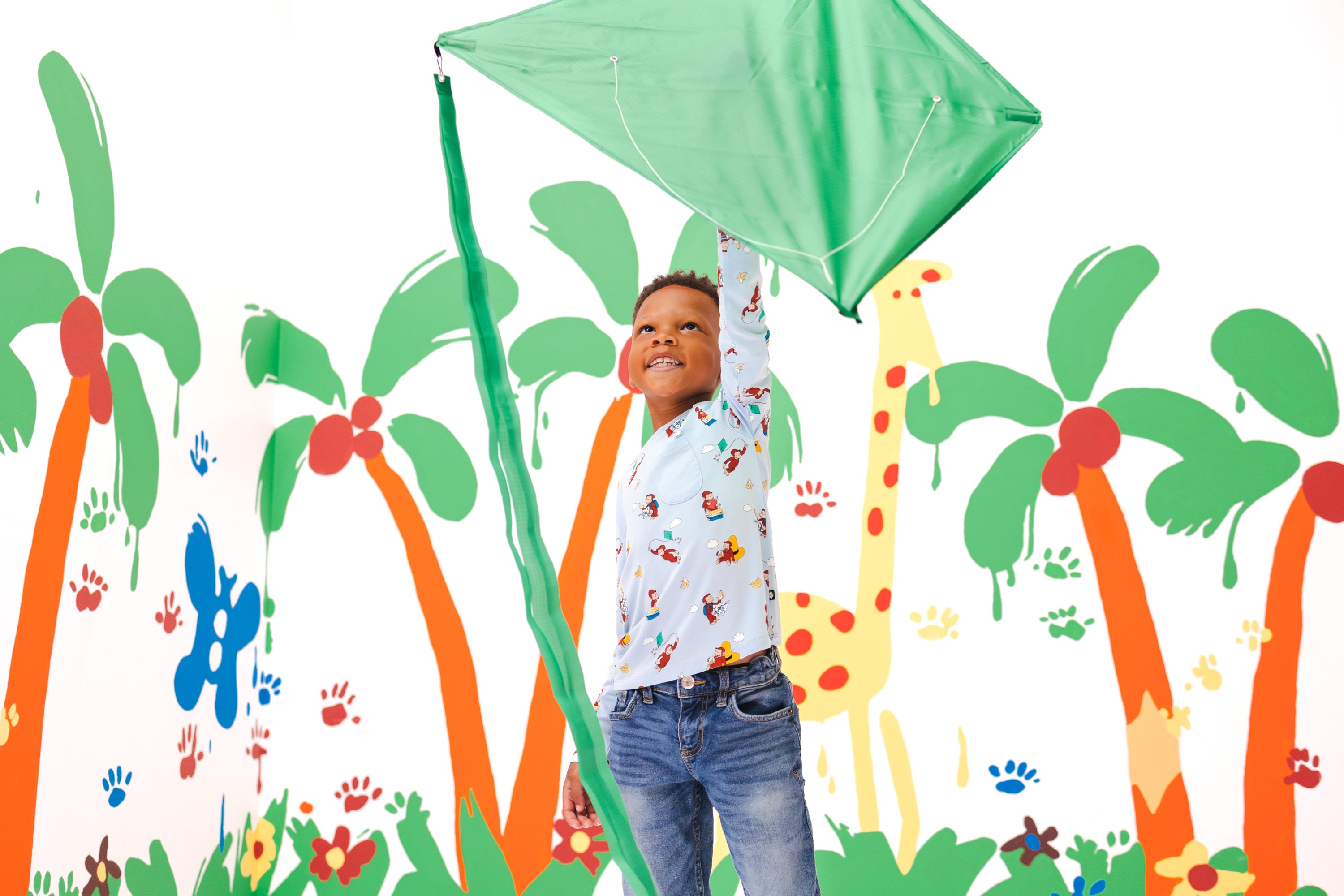 A young boy wearing a white patterned long-sleeve curious george shirt and blue jeans stands holding up a green diamond-shaped kite against a white wall decorated with colorful cartoon palm trees, flowers, and a giraffe.