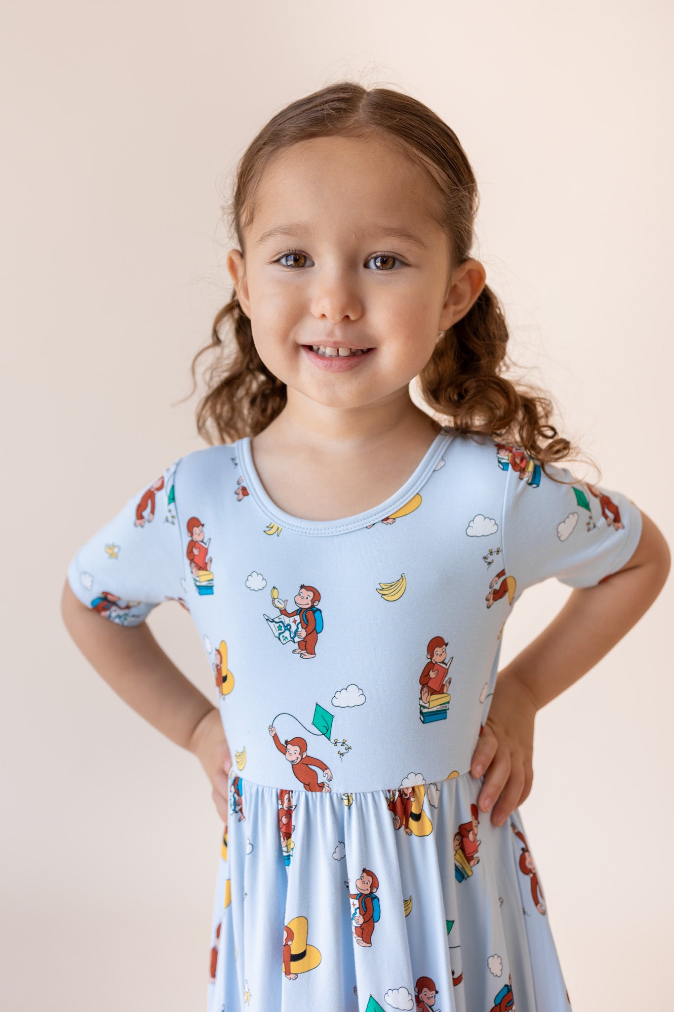 A young girl with brown hair in pigtails smiles while wearing a light blue short-sleeve dress with a colorful cartoon print, posed against a solid light beige background.