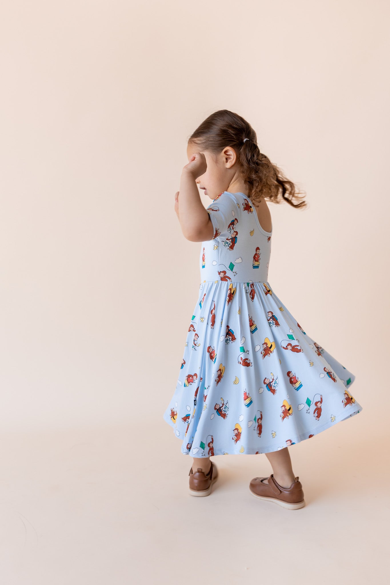 A young girl with brown hair in a ponytail is shown from behind wearing a light blue sleeveless dress with a colorful cartoon animal print, standing against a solid beige studio background.