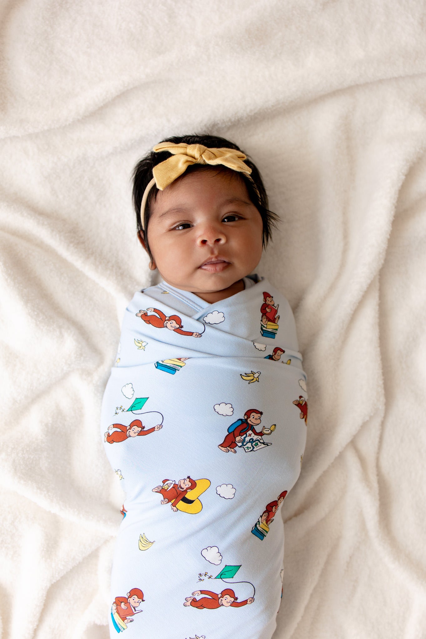 Baby swaddled in a Blue Swaddle Blanket with Curious George print lying on a beige sheet