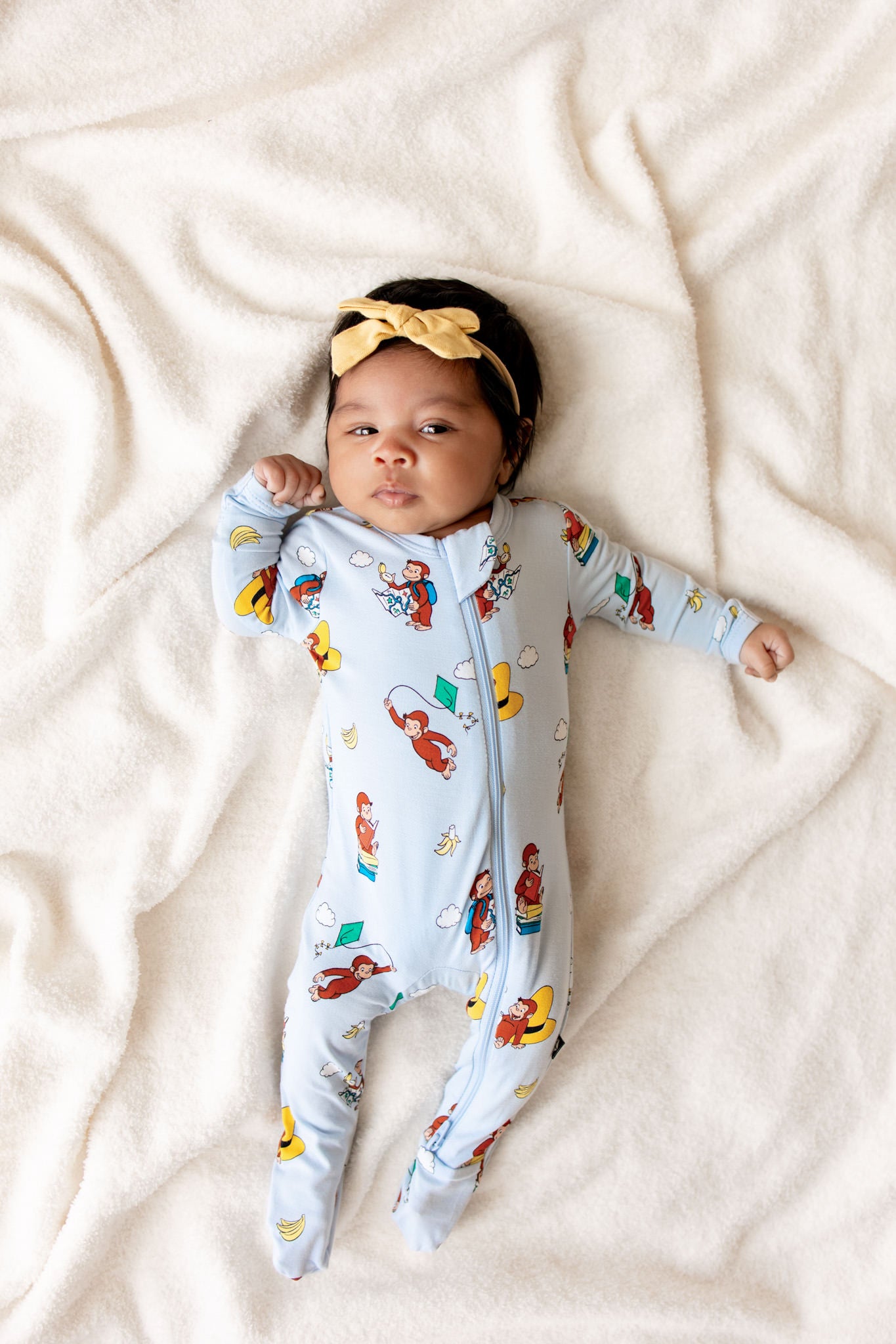 Baby wearing Blue Zippered Footie in Curious George print lying on beige sheet