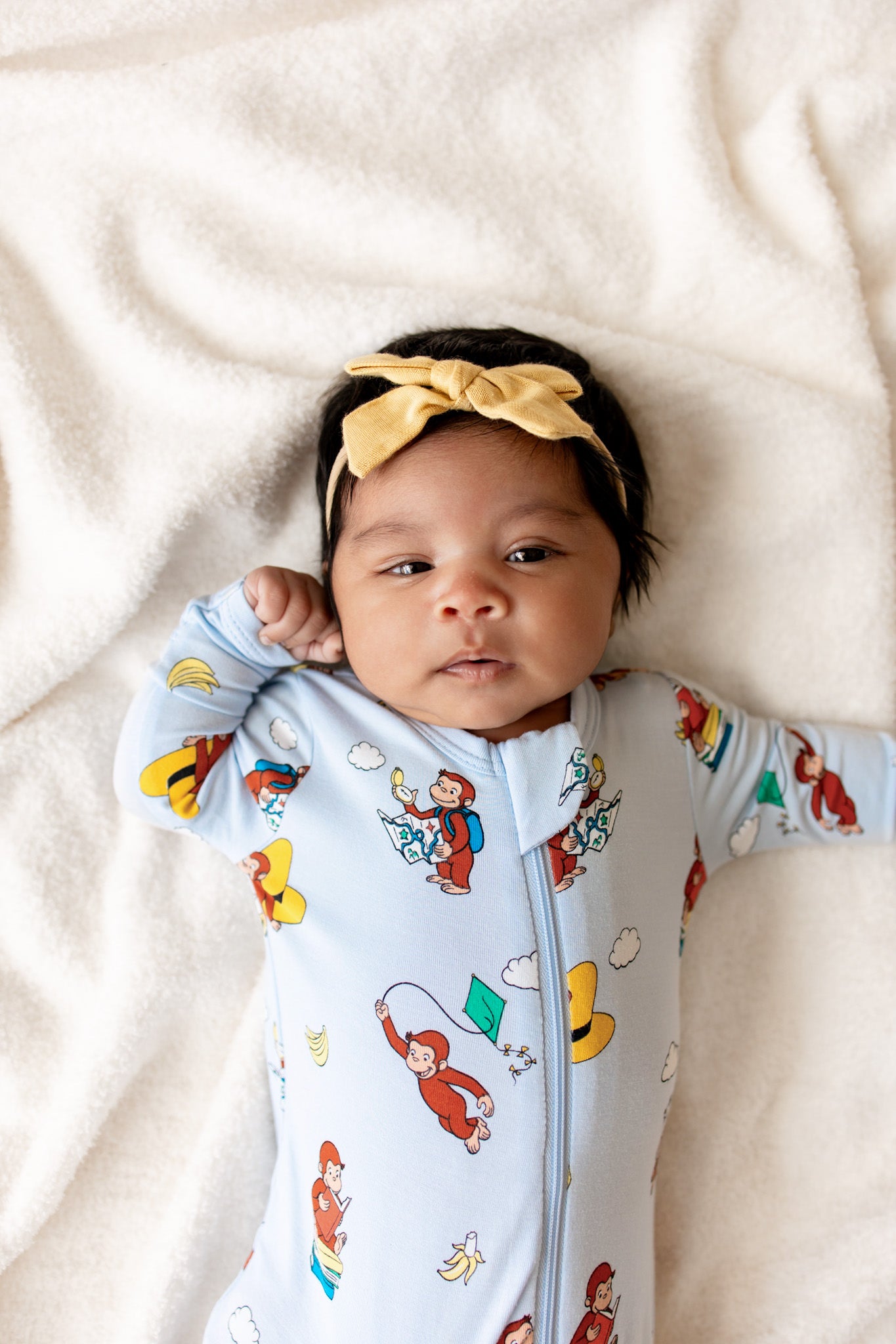 Baby wearing Blue Zippered Footie in Curious George print lying on beige sheet