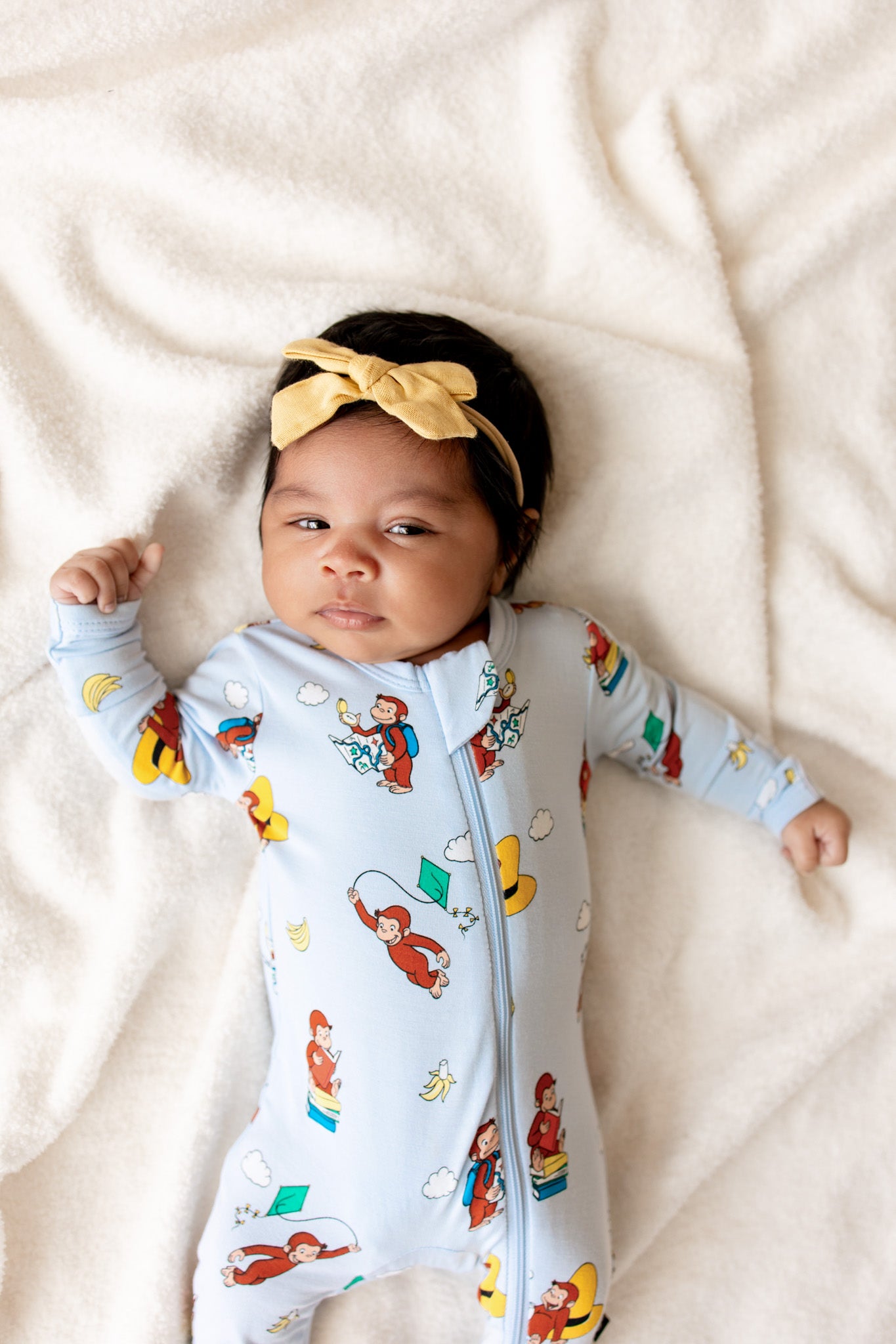 Baby in Blue Zippered Footie in Curious George print lying on beige sheet
