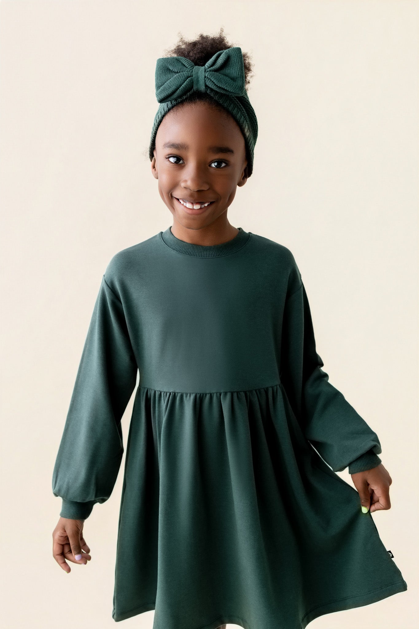 Girl wearing Long Sleeve Skater Dress in Cypress color with chunky knit knotted bow headband on a beige background