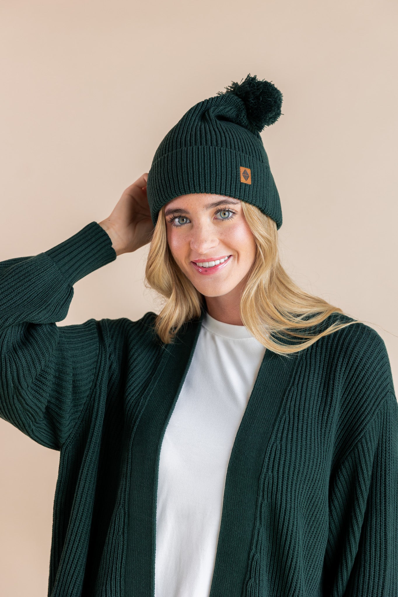 Modal smiling in chunky knit adult ribbed beanie and chunky knit adult cardigan in cypress over white t-shirt on a beige background