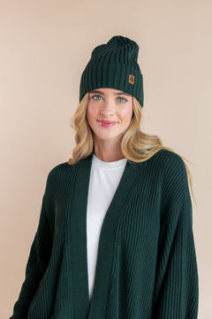 Modal smiling in chunky knit women's oversized cardigan and chunky knit adult ribbed beanie in cypress over white t-shirt on a beige background