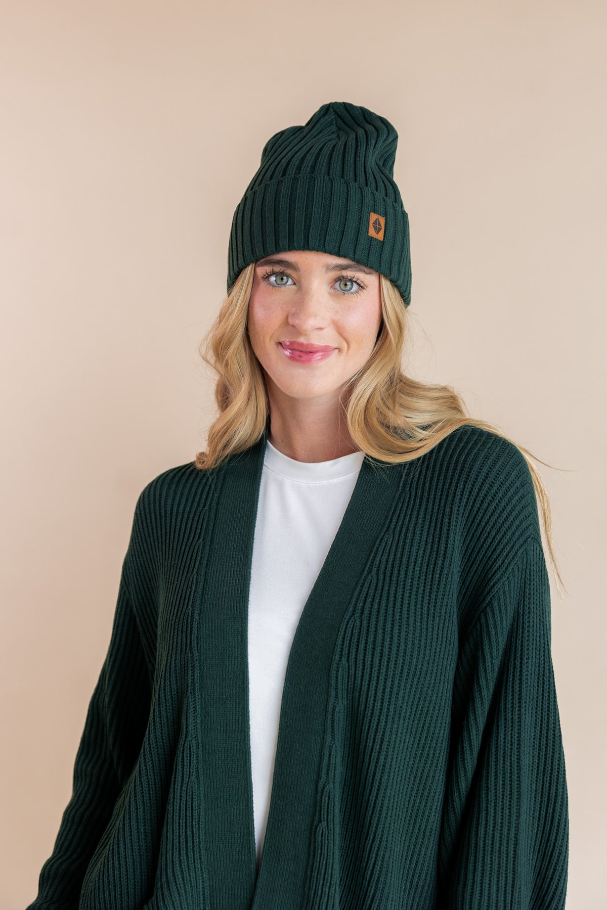 Modal smiling in chunky knit women's oversized cardigan and chunky knit adult ribbed beanie in cypress over white t-shirt on a beige background