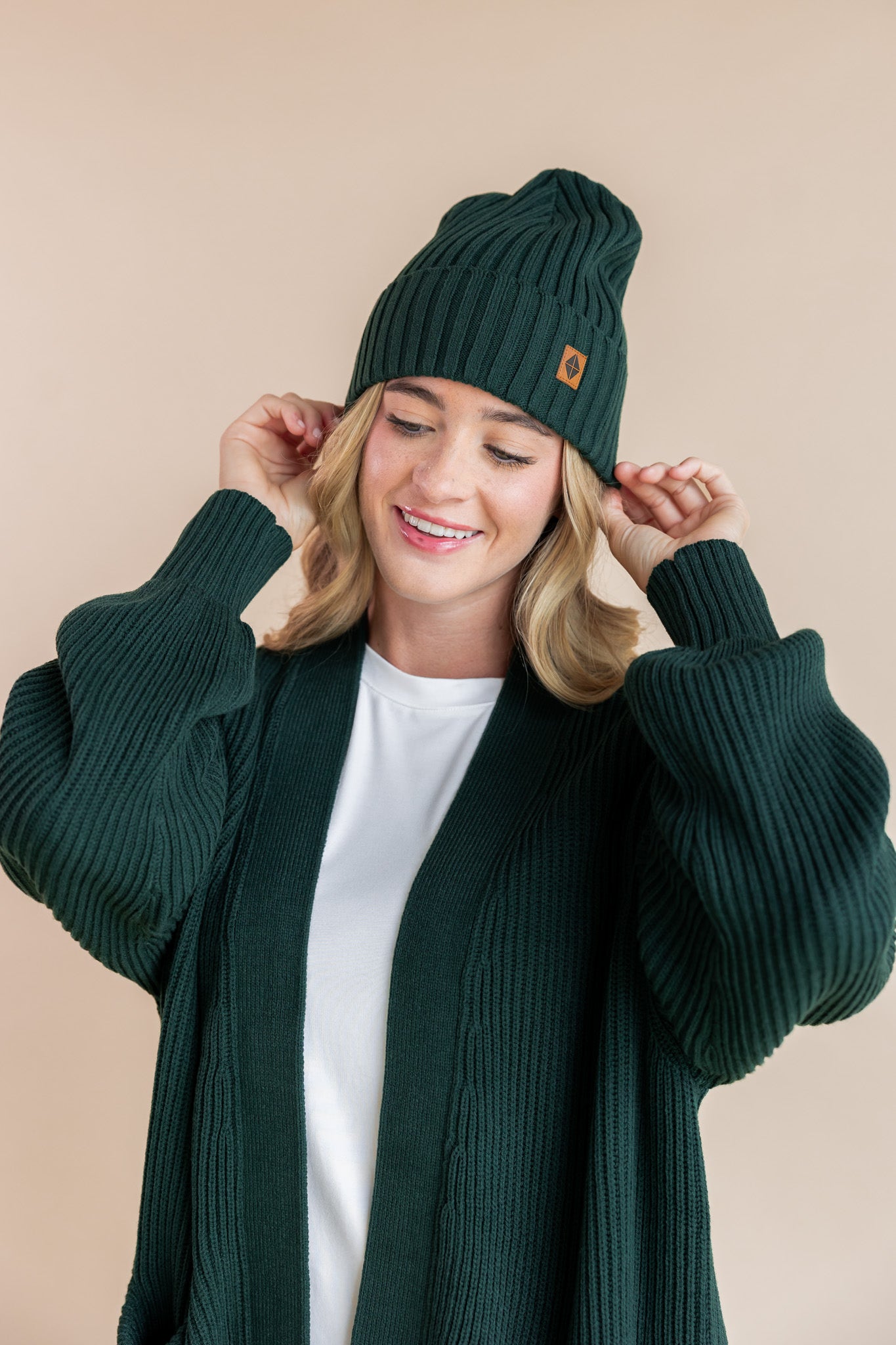 Modal posing in chunky knit women's oversized cardigan and chunky knit adult ribbed beanie in cypress on a beige background