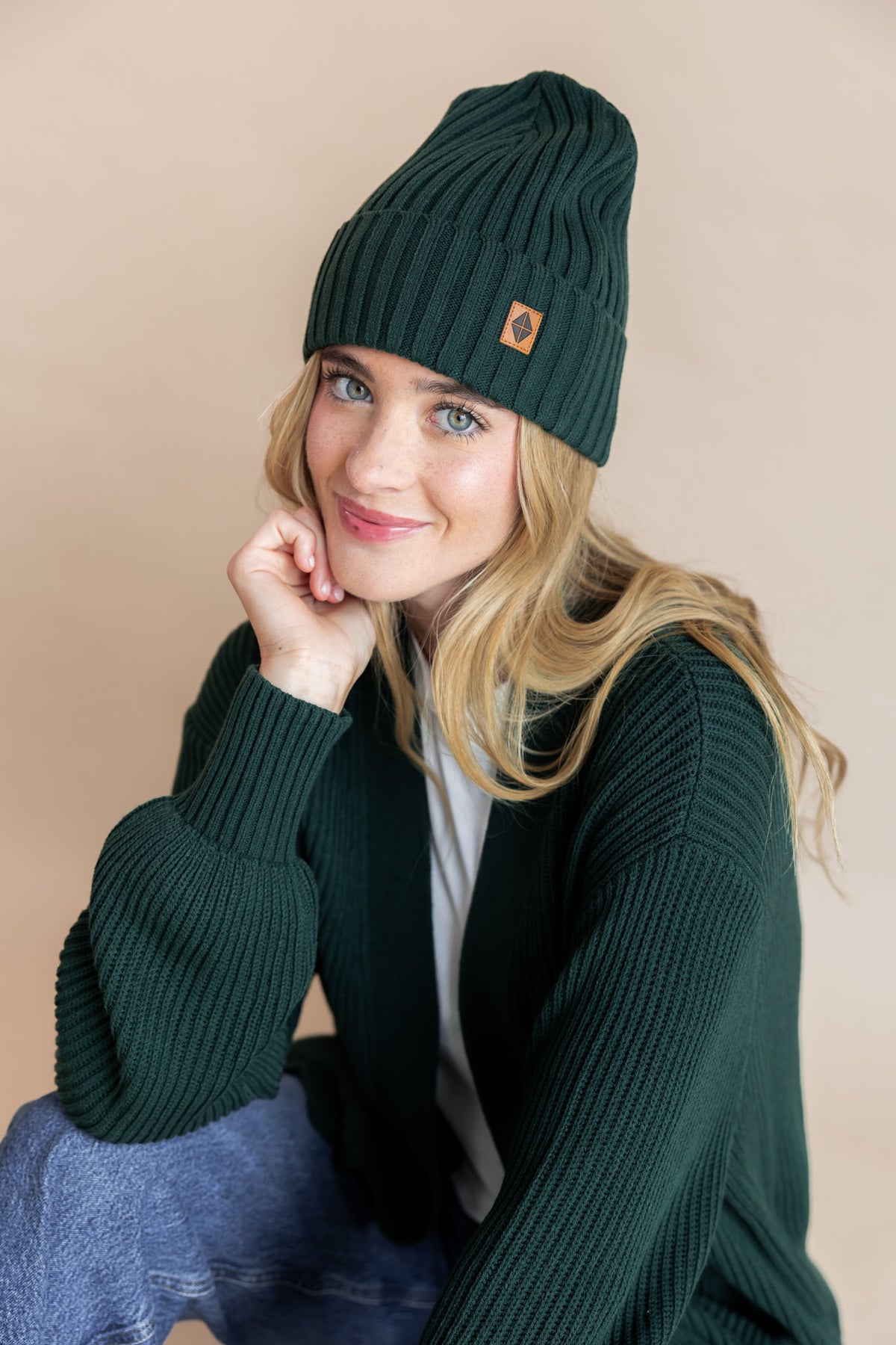 Modal smiling in chunky knit women's oversized cardigan and chunky knit adult ribbed beanie in cypress over blue jeans on a beige background