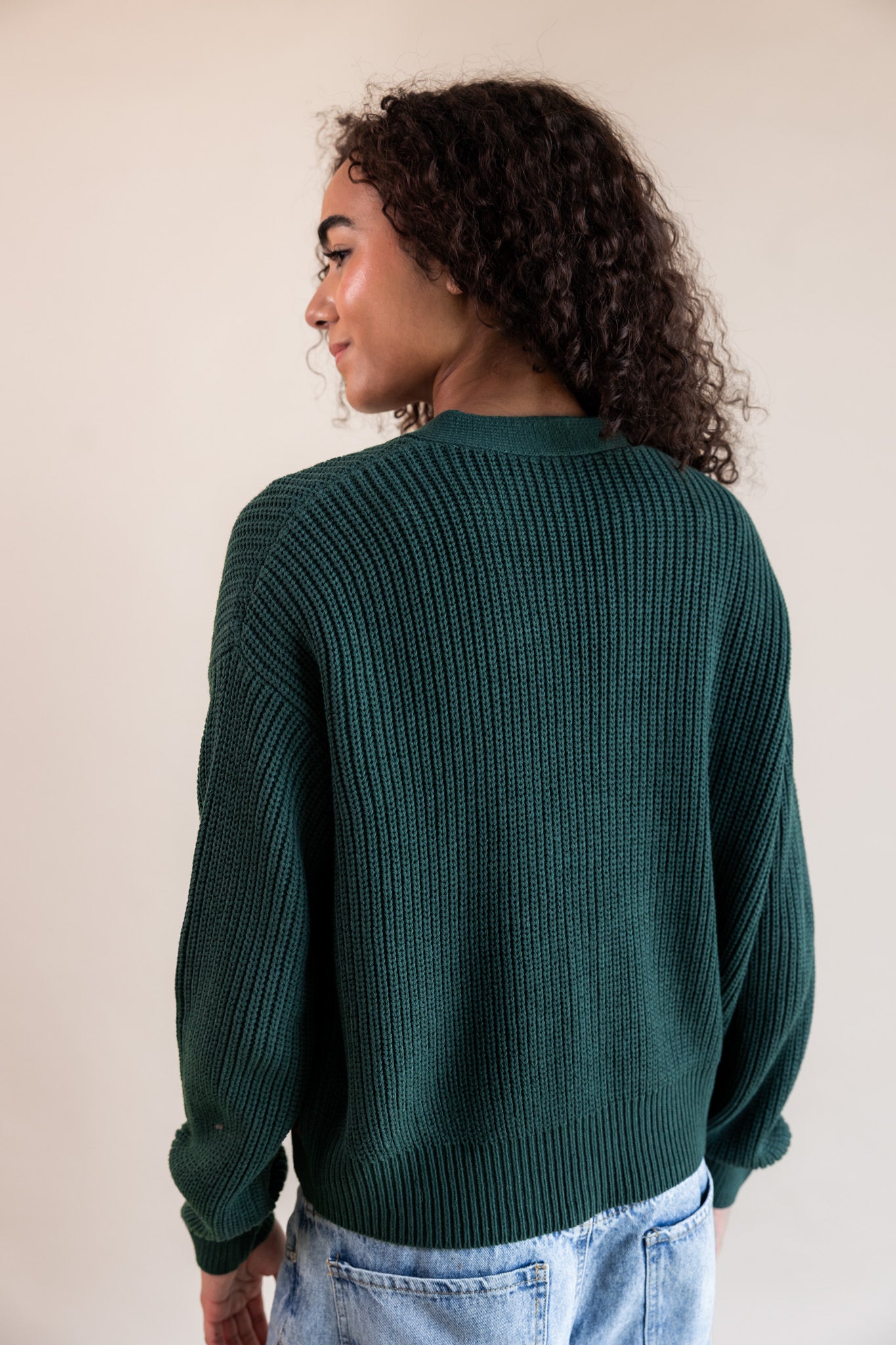 Modal showcasing the back side of chunky knit cropped cardigan in cypress over blue jeans on a beige background
