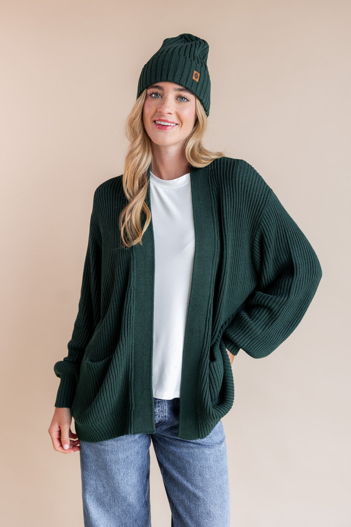 Modal wearing chunky knit women's oversized cardigan and chunky knit adult ribbed beanie in cypress over blue jeans on a beige background