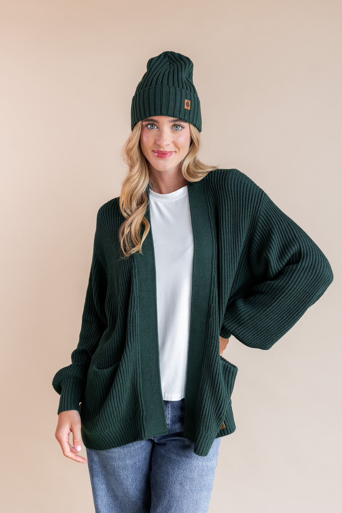 Modal wearing chunky knit women's oversized cardigan and chunky knit adult ribbed beanie in cypress over blue jeans on a beige background