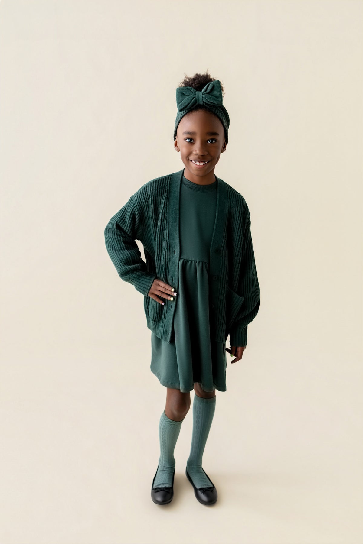 Girl wearing Chunky Knit Oversized Cardigan in Cypress color over similar color Long sleeve skater dress and Chunky knit knotted bow headband and knee high socks on a beige background