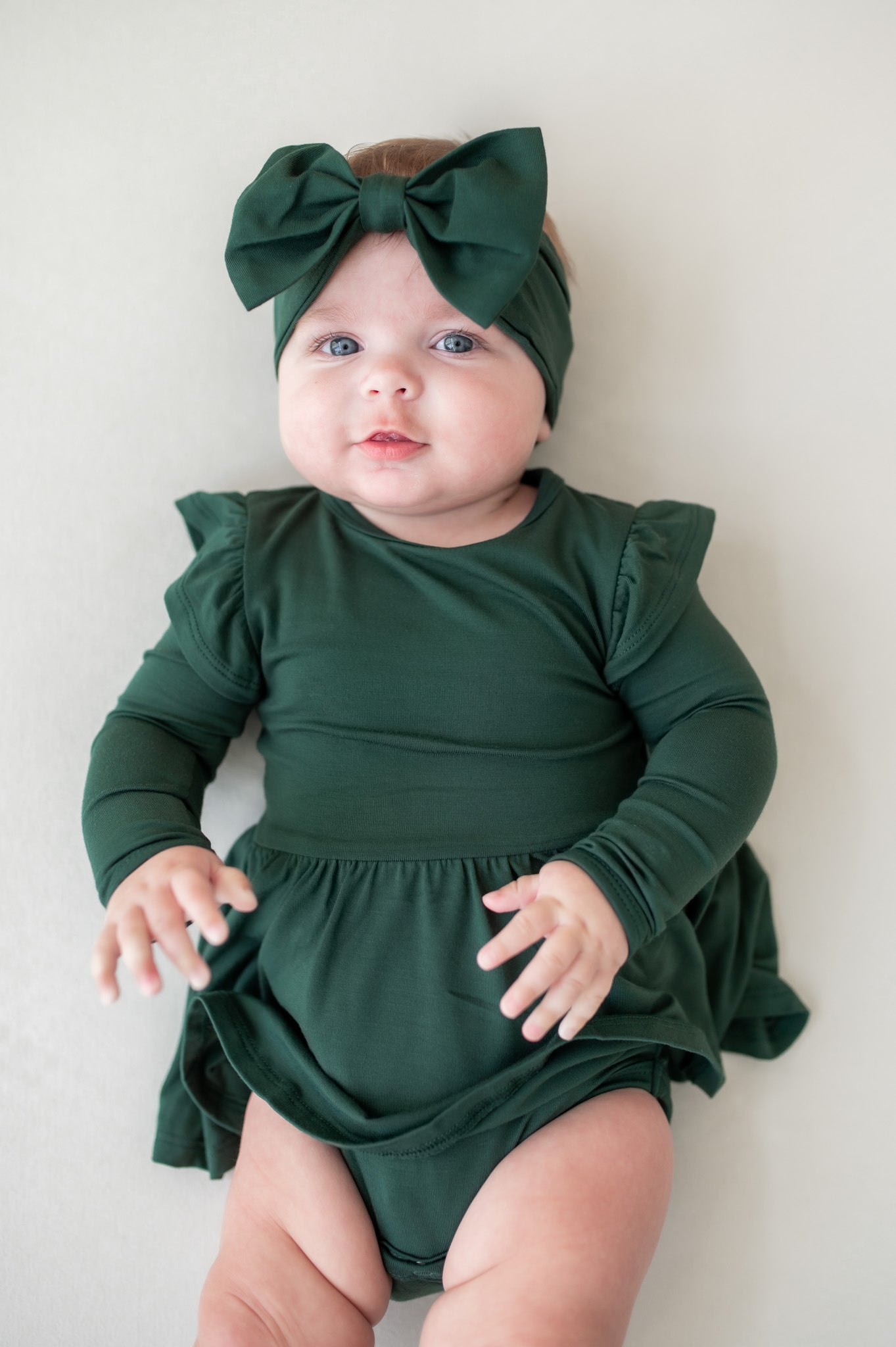 Baby wearing Twirl bodysuit dress in cypress with knotted bow headband in cypress on a beige background