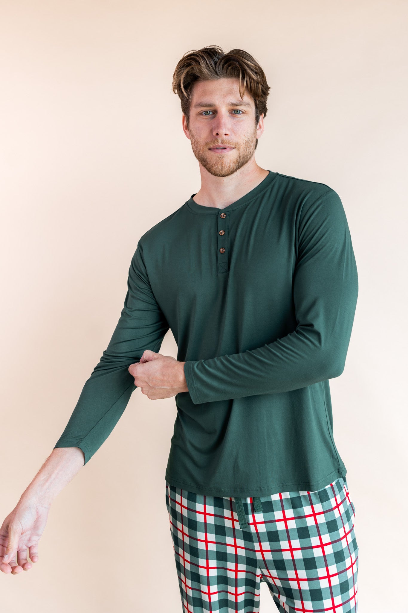Man wearing long sleeve henley tee in cypress color over men's lounge pants in holiday plaid on a beige background