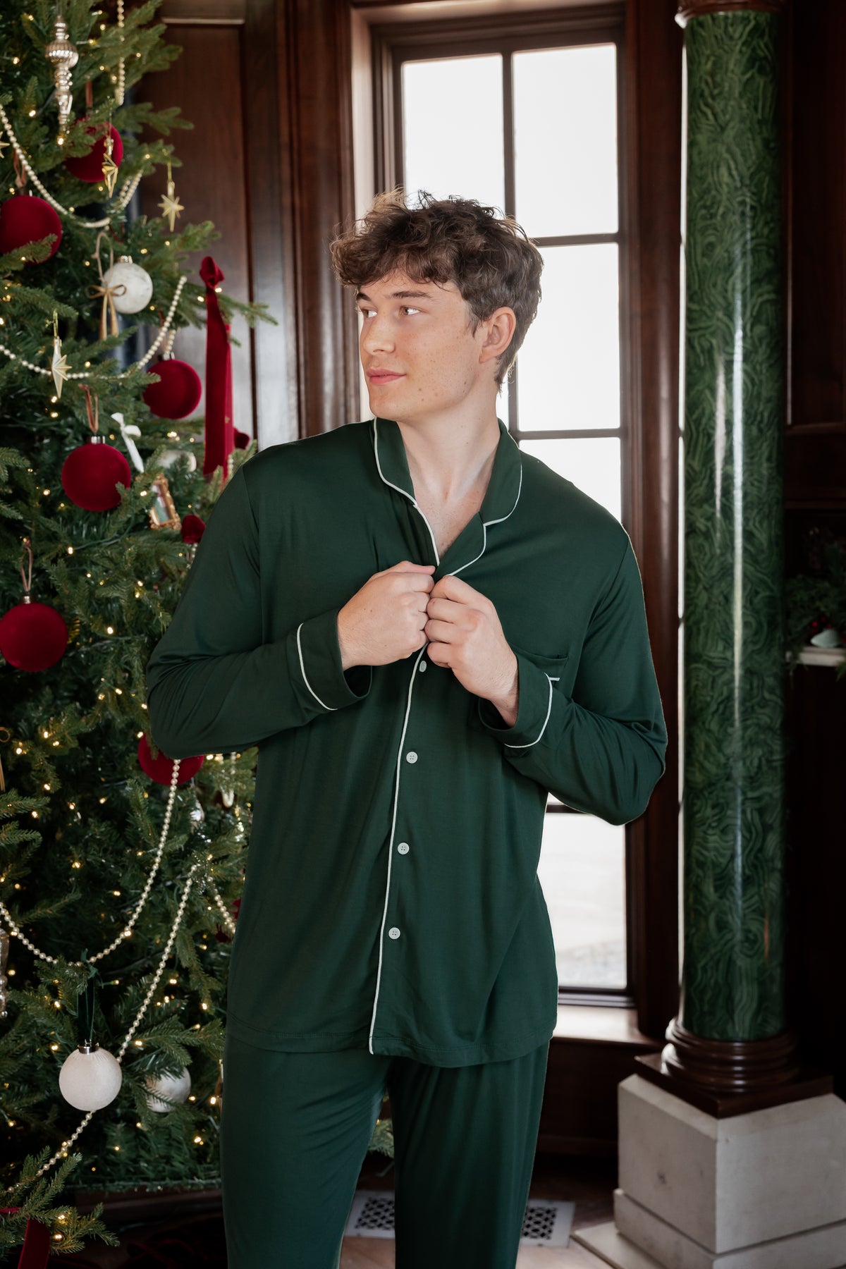 A man wearing a dark green long-sleeve pajama set with white piping stands in front of a window and a decorated Christmas tree.