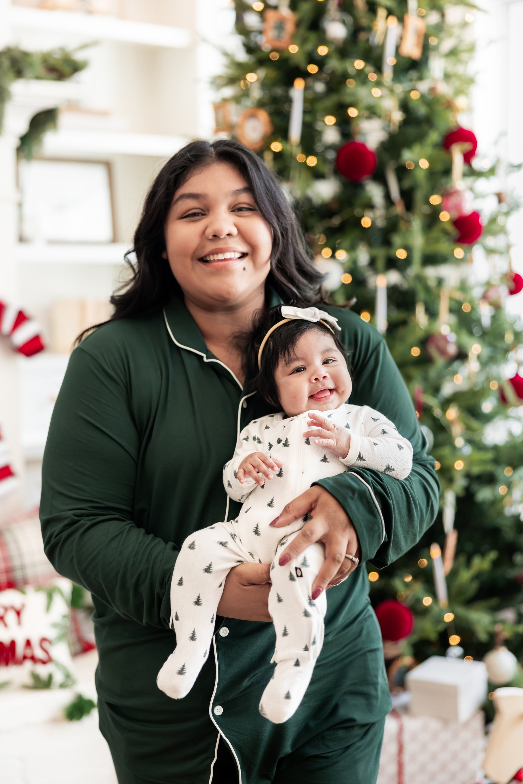 Mommy wearing long sleeve pajama set in cypress holding her Baby wearing Pointelle Zippered Footie in Christmas Trees with a Christmas tree in the background