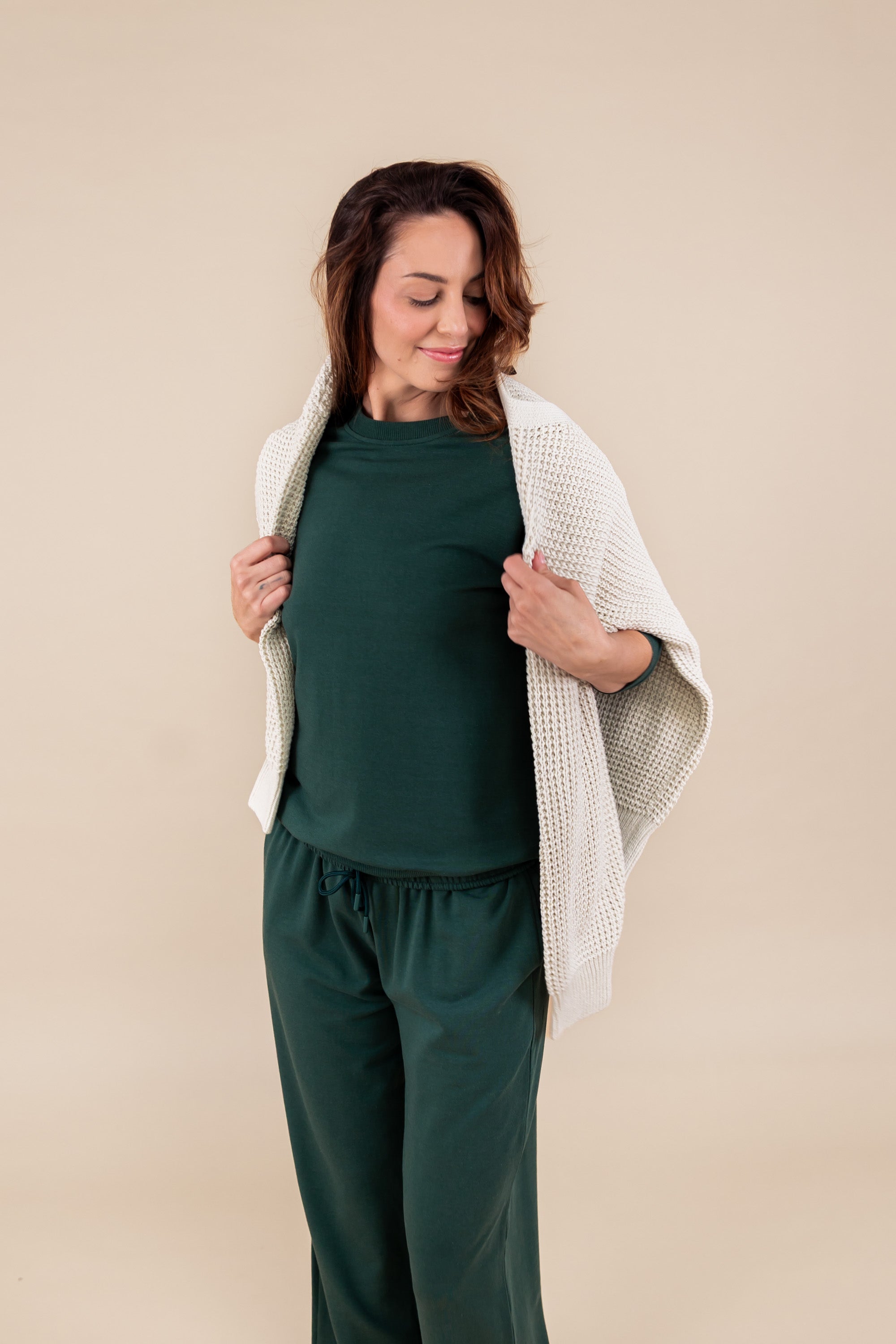Woman wearing Women's Sweatpants in Cypress color with beige color sweater over her shoulders under similar color women's sweatshirt on a beige background