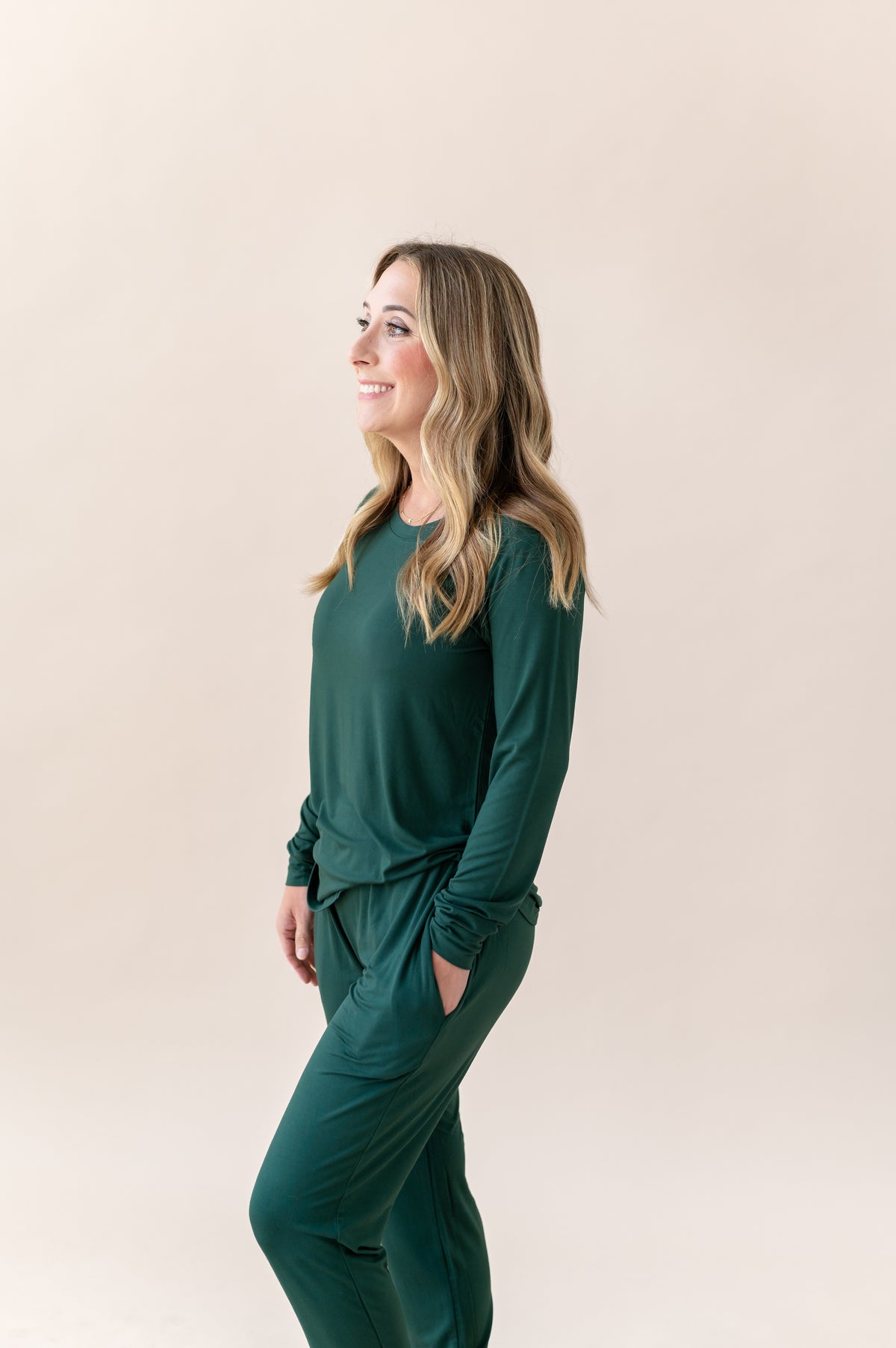 A woman wearing a matching dark green long-sleeve top and jogger pants set, standing in profile against a solid light beige background.