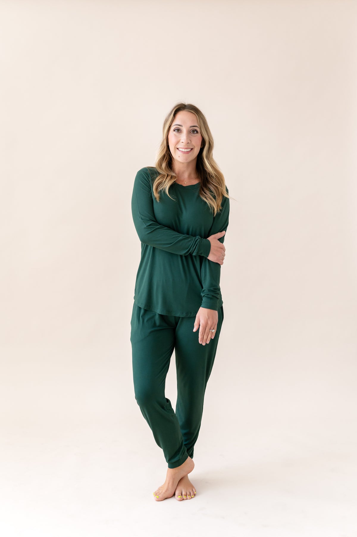 A woman modeling a dark green long-sleeve pajama set consisting of a crewneck top and matching pants, standing against a solid light beige background.