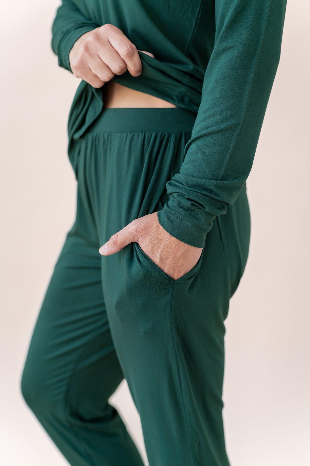 A close-up of a person wearing a dark green matching lounge set, featuring a long-sleeve top with a side cutout and matching drawstring-free pants with side pockets.