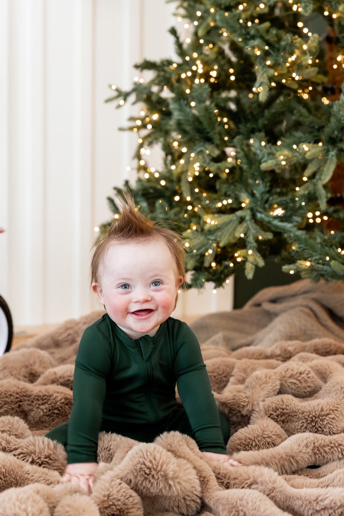 A baby wearing a dark green long-sleeve bodysuit sits on a fluffy tan blanket in front of a lit Christmas tree.