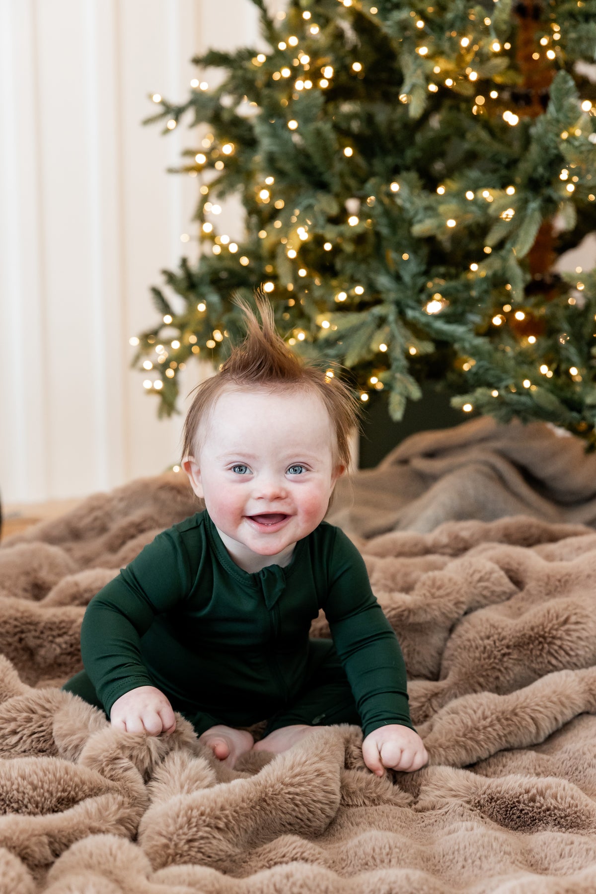 A baby wearing a dark green long-sleeve romper sits on a brown faux fur blanket in front of a lit Christmas tree.