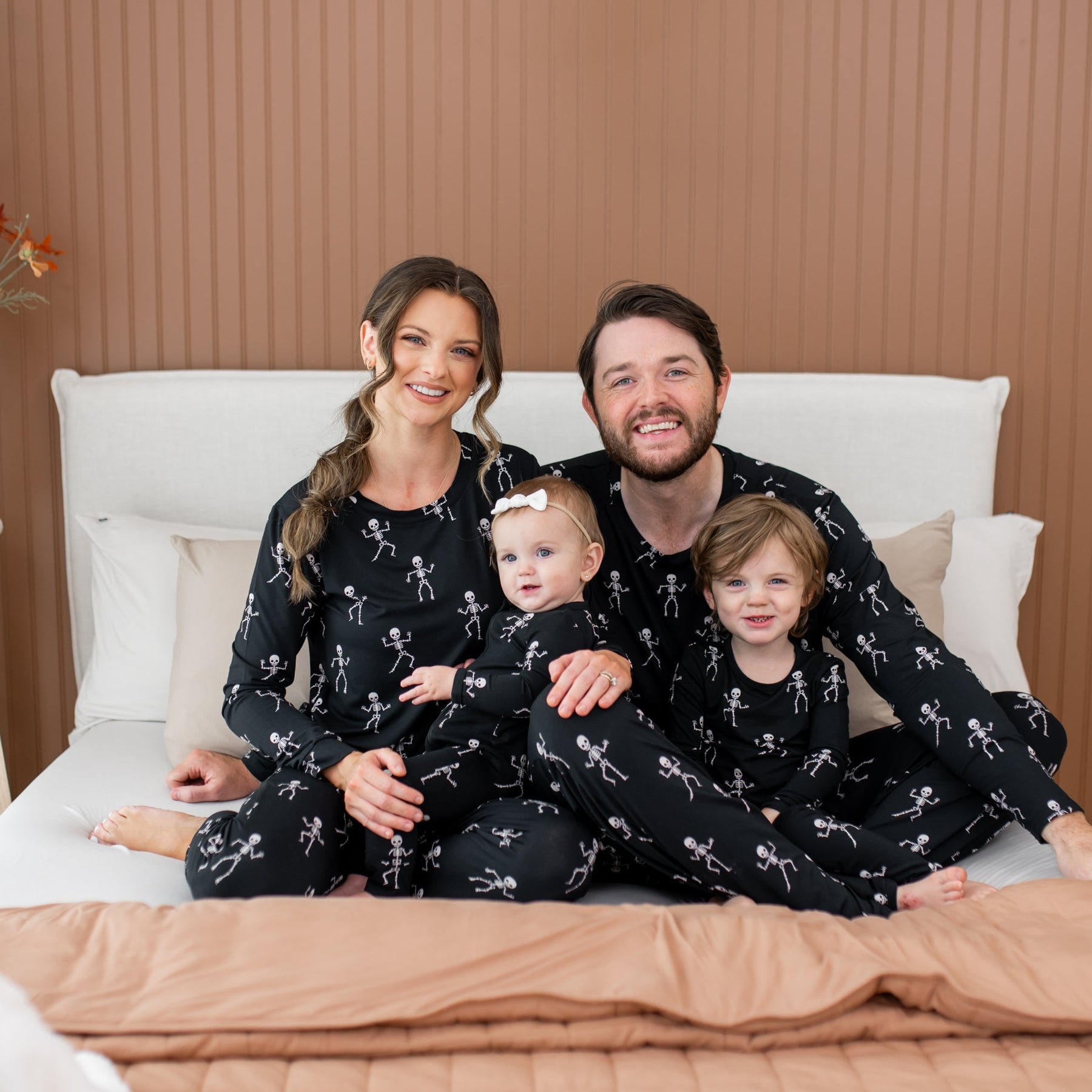 Family of four wearing matching halloween pajamas with a skeleton print sitting on a bed in a cozy bedroom.