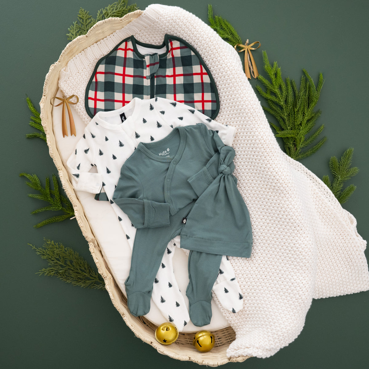 A baby clothing set arranged in a woven basket with a white knit blanket, green pine branches, and small gold bells on a dark green background. The set includes a plaid bib, a white long-sleeve top with small christmas tree prints, a dusty sage green long-sleeve top with a front knot detail, matching green pants