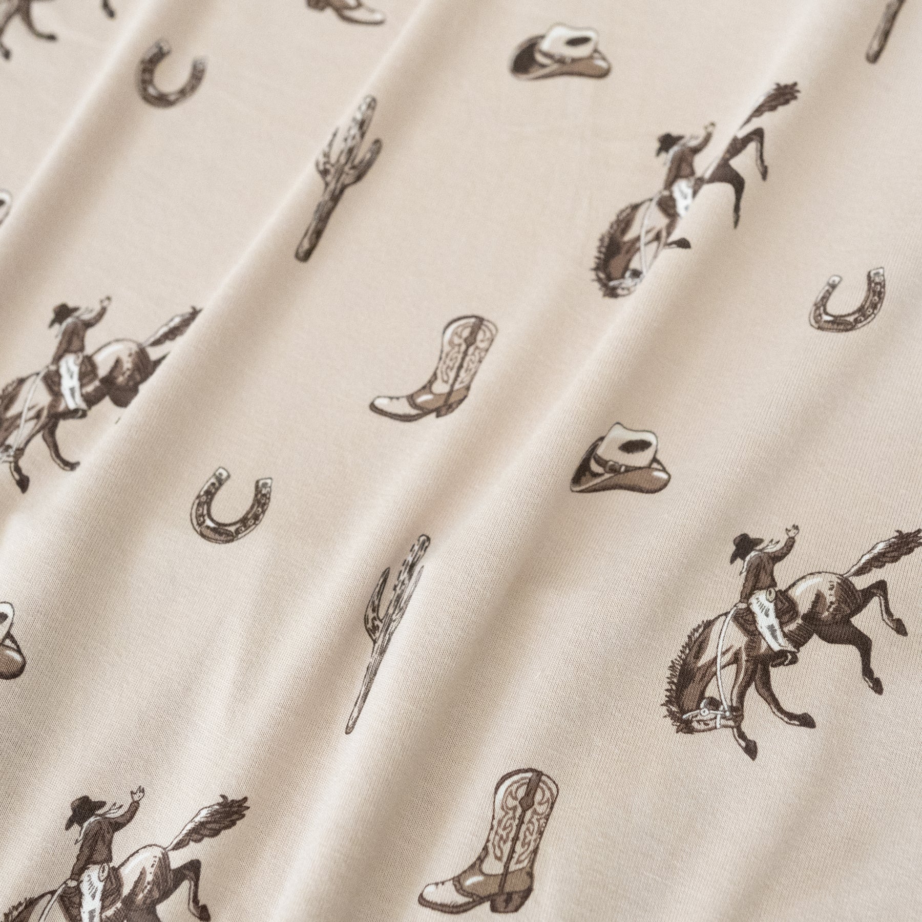 Close up of bamboo fabric: A repeating, all-over print in monochromatic browns, featuring: a cowboy riding a bucking horse; a western boot: a three-arm cactus; a horseshoe; and a cowboy hat, over a dusty beige color named Bisque.