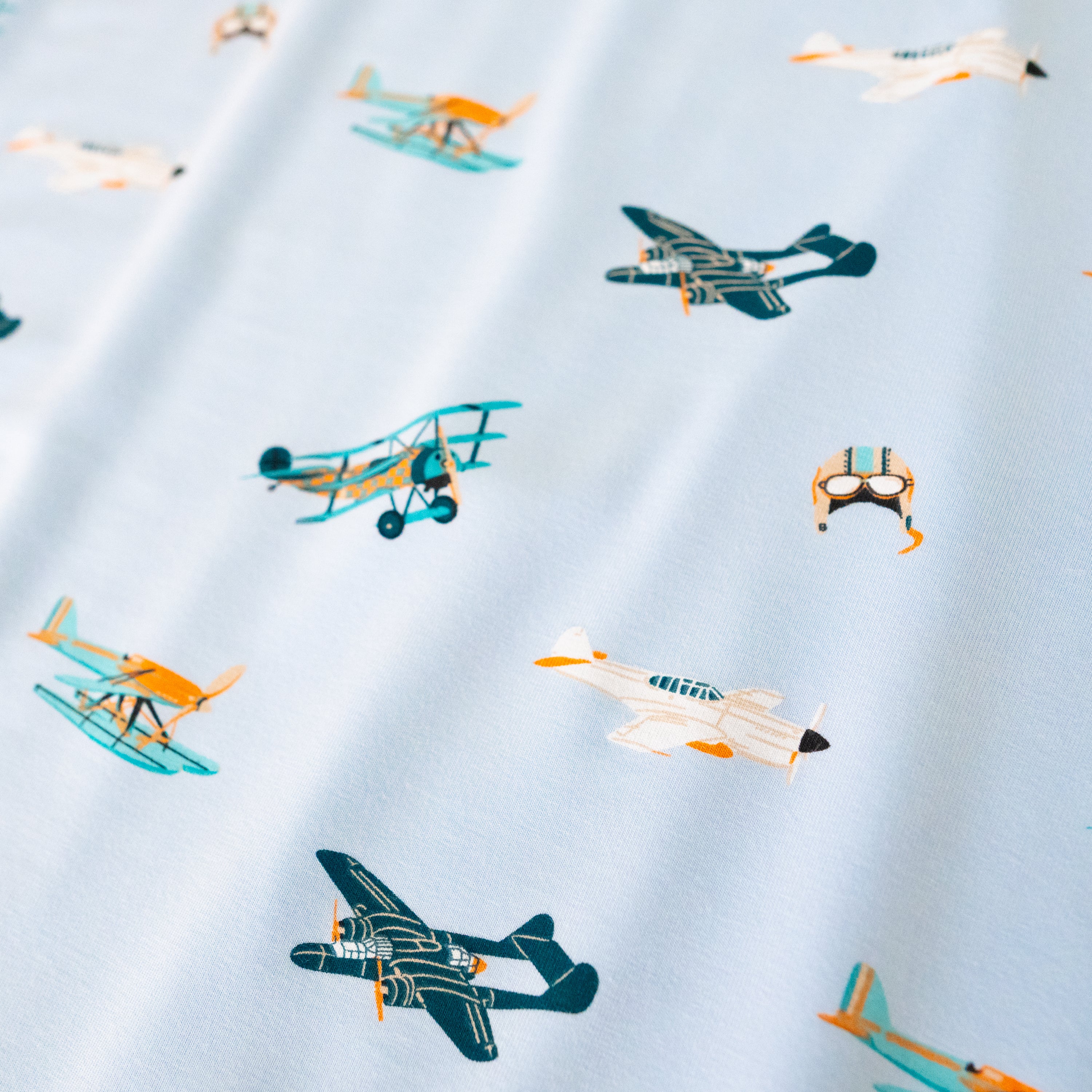 A repeating, all-over print featuring: a teal triplane with a light orange checker design; a racing seaplane colored in turquoise and light orange; a night fighter aircraft in teal with light orange accents; a white fighter bomber aircraft with a black nose, teal windows, and light orange accents; and a light grey aviator hat with goggles and blue stripes, over a sky blue background color called Breeze.