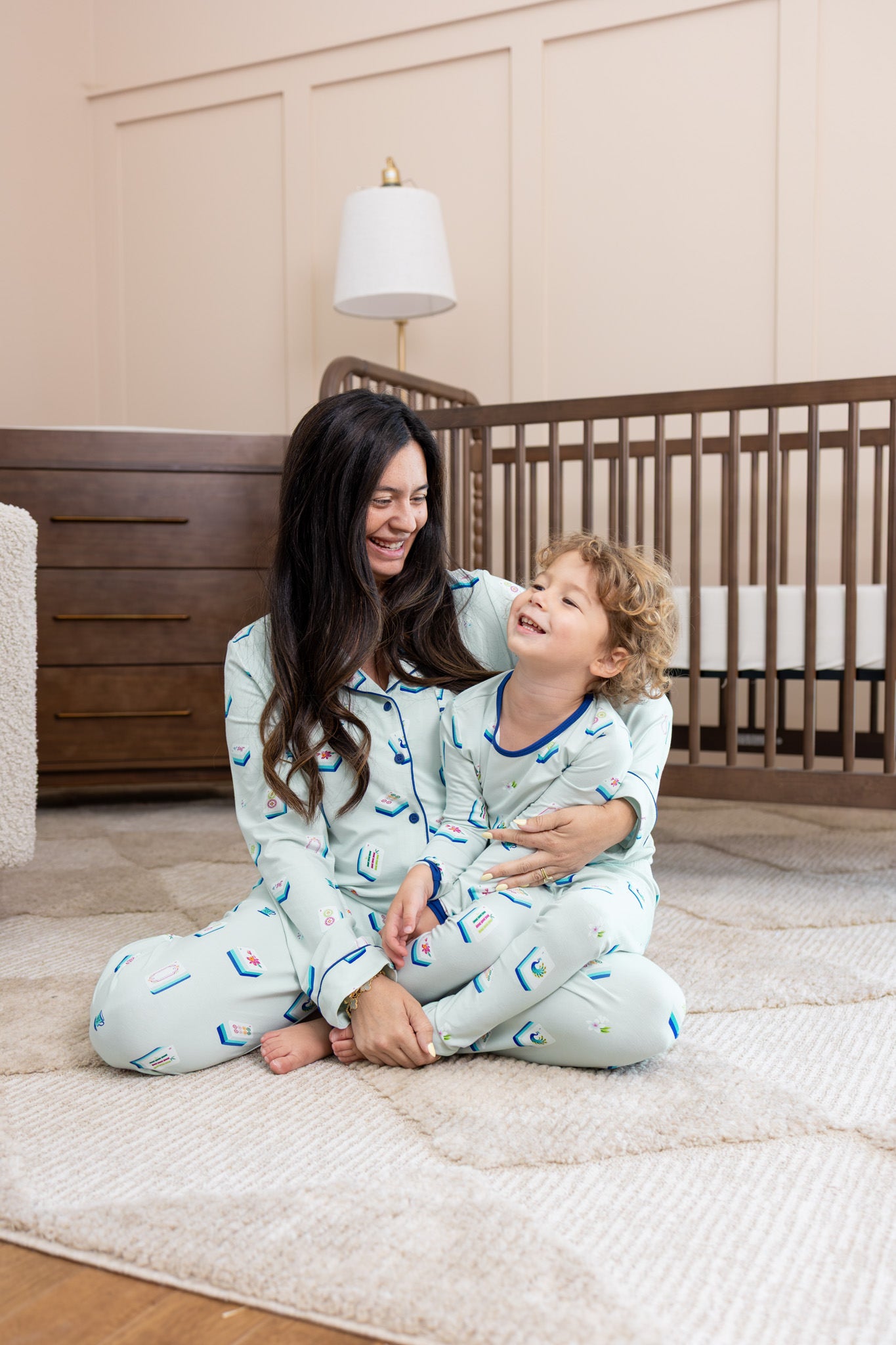 A woman and a young boy wearing matching light blue pajamas with a small blue mahjong pattern, sitting together on a cream rug in a nursery.