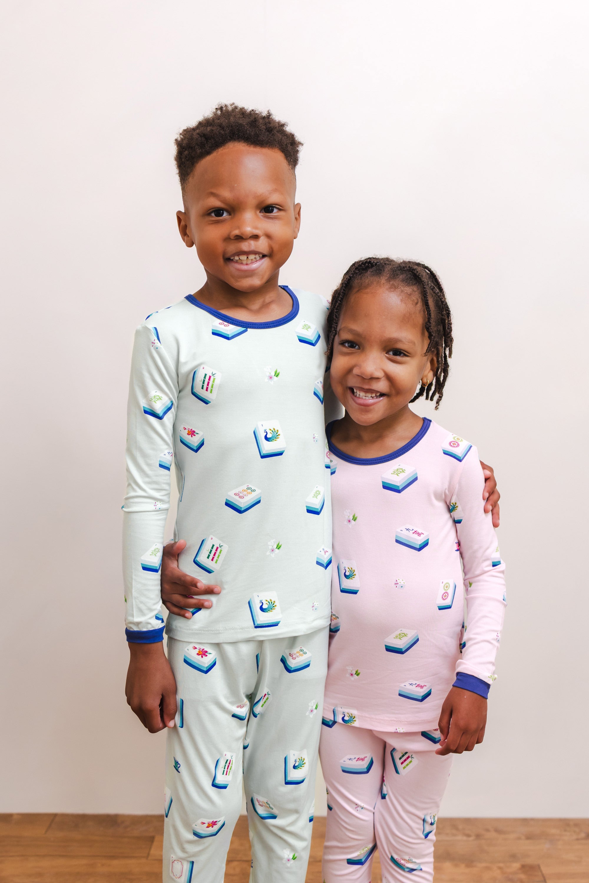 Boy and Girl standing together wearing Long Sleeve Pajamas in Dew Mahjong print and Long Sleeve Pajamas in Sakura Mahjong print against a beige background