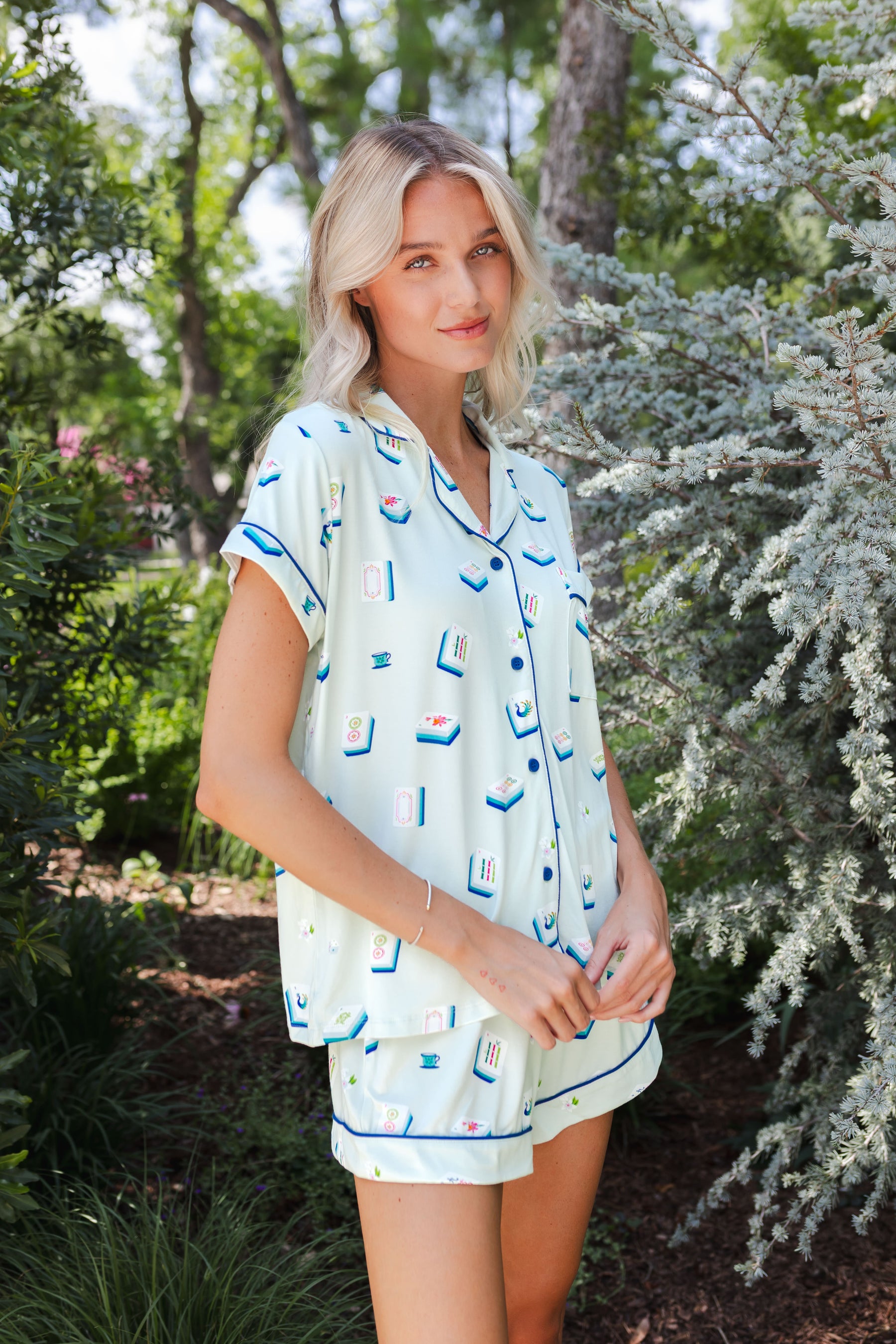 woman wearing light green short sleeve pajama set with mahjong tile print standing in front of greenery 