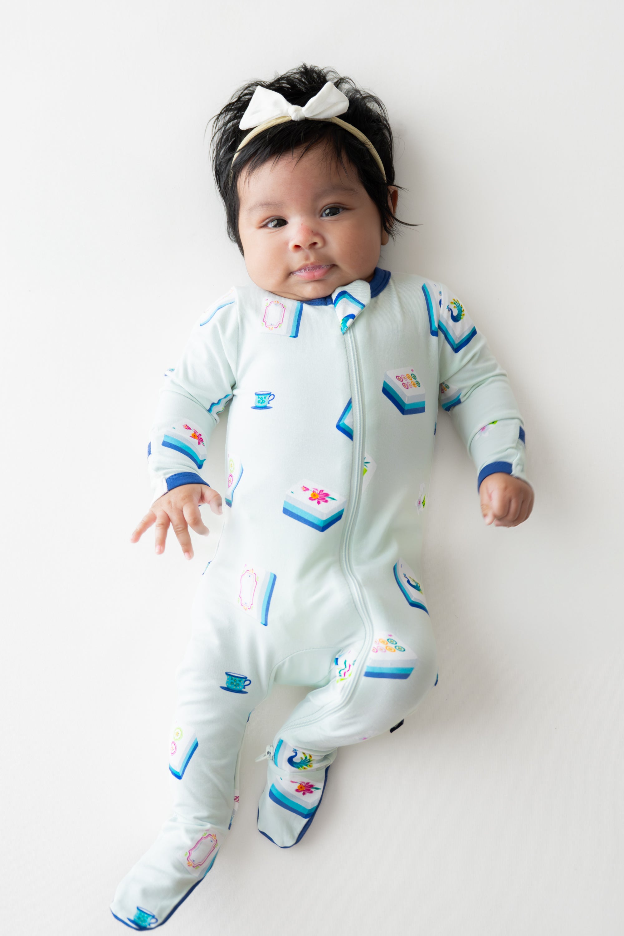 Baby wearing Zippered Footie in Dew Mahjong print and a bow headband on a beige background