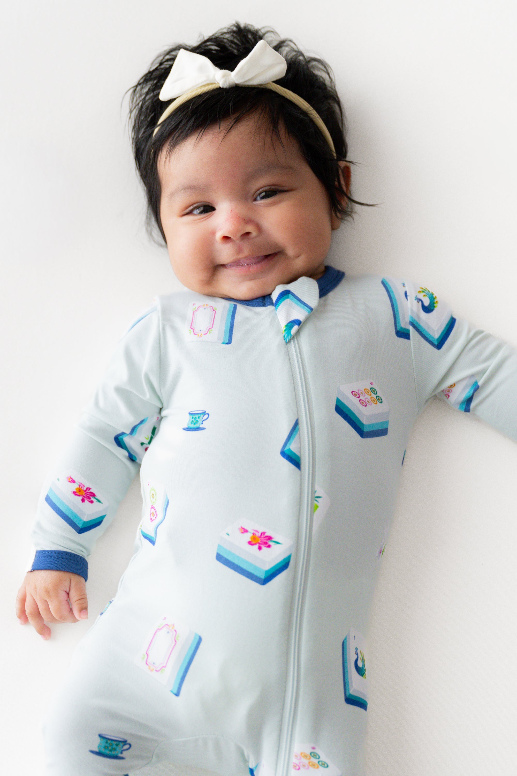 Baby smiling in Zippered Footie in Dew Mahjong print and a headband on a beige background