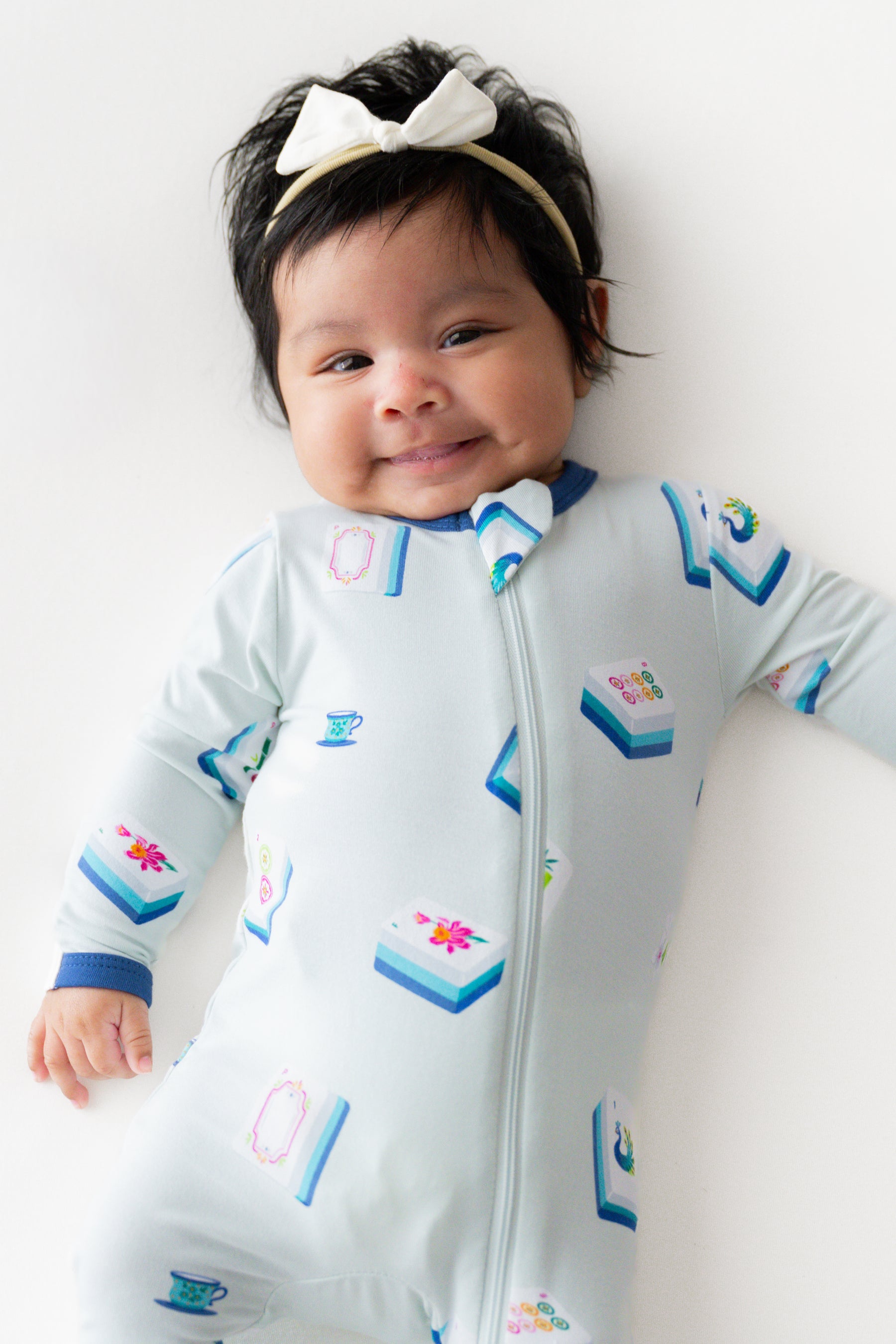 Baby smiling in Zippered Footie in Dew Mahjong print and a headband on a beige background