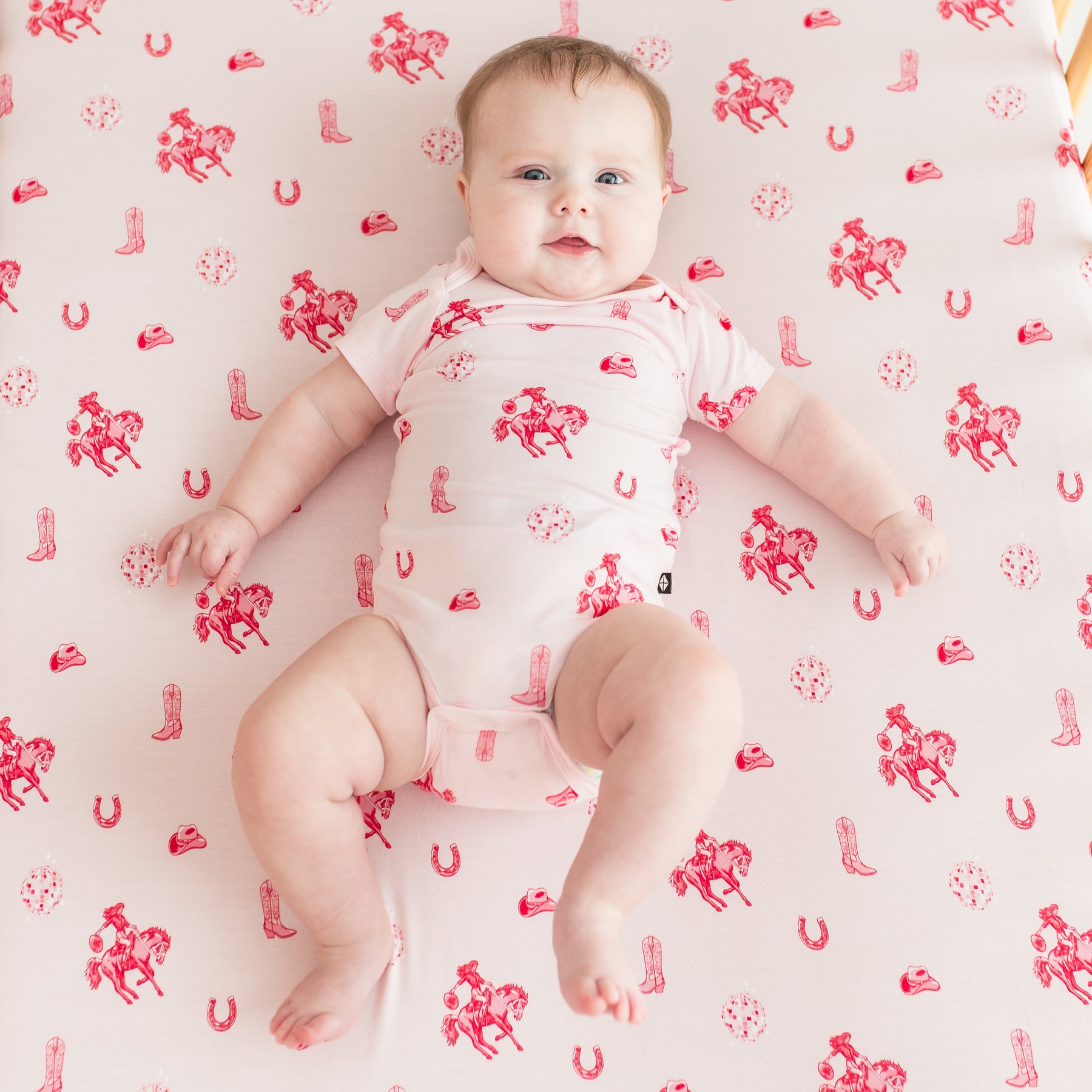 Infant girl wearing the Bodysuit in Disco Cowgirl laying in a crib on a matching crib sheet