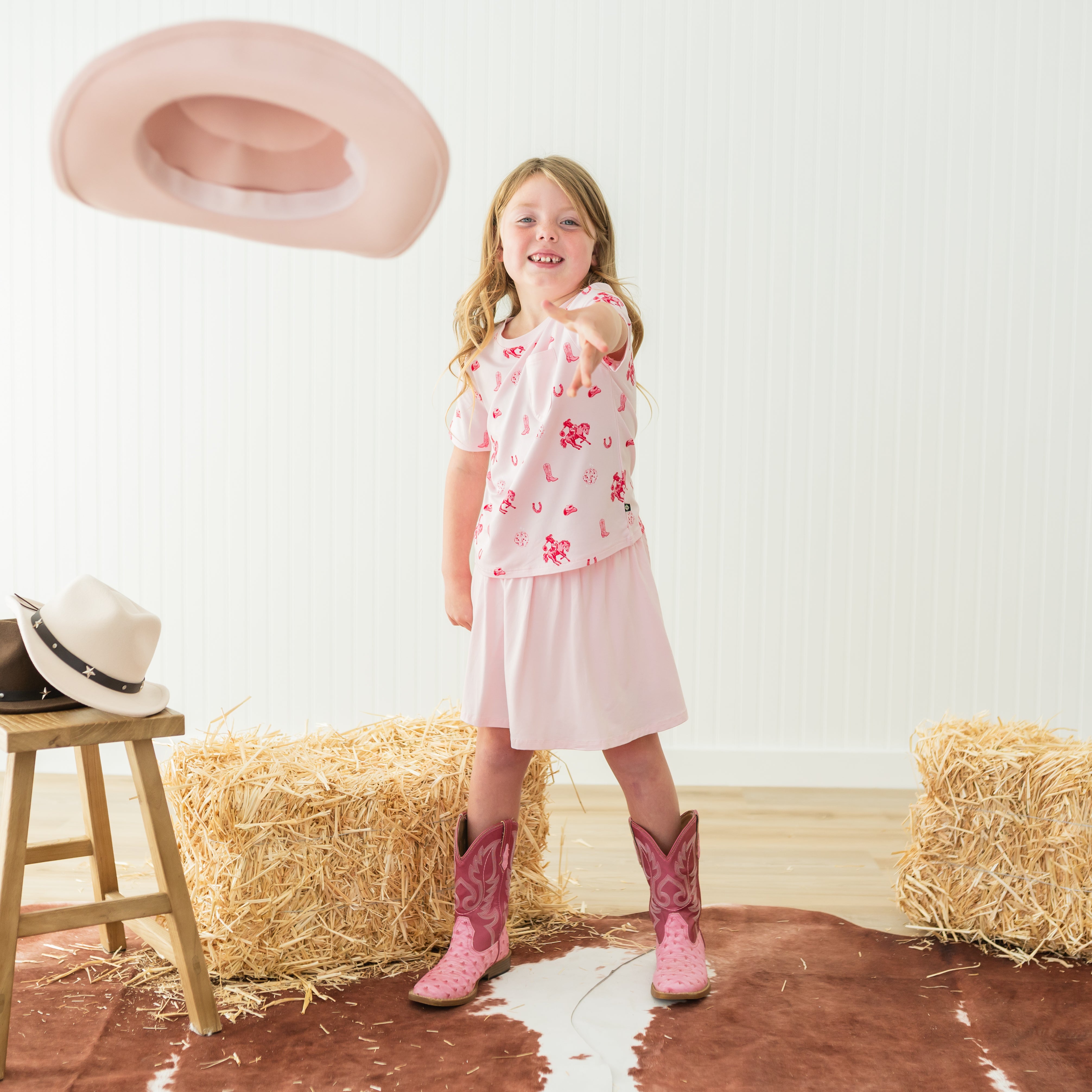 Young girl standing wearing the Toddler Crew Neck Tee in Disco Cowgirl with a pink skirt throwing a pink cowboy hat