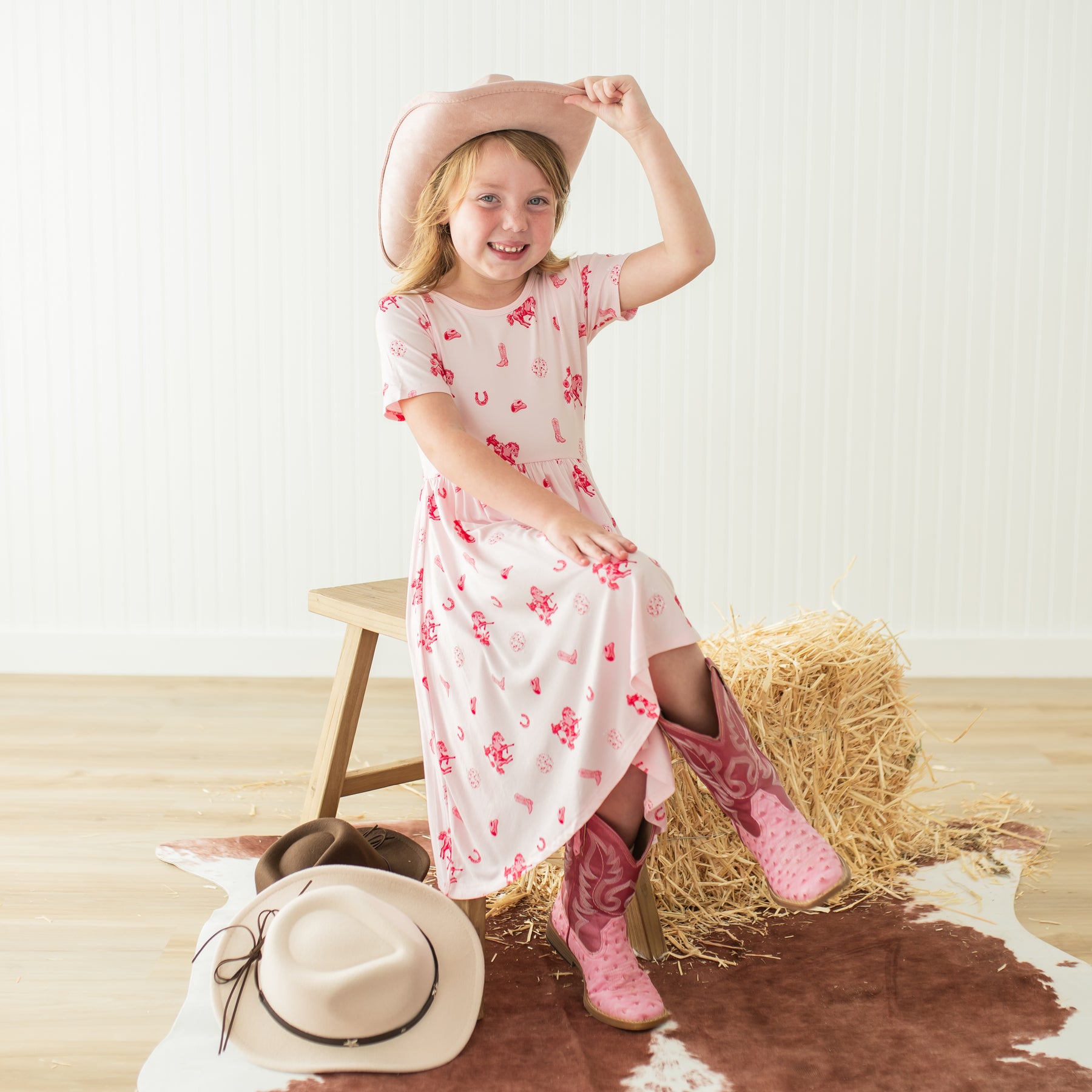 Young girl sitting on a wooden bench wearing the Twirl Dress in Disco Cowgirl with a pink cowboy hat and pink cowboy boots