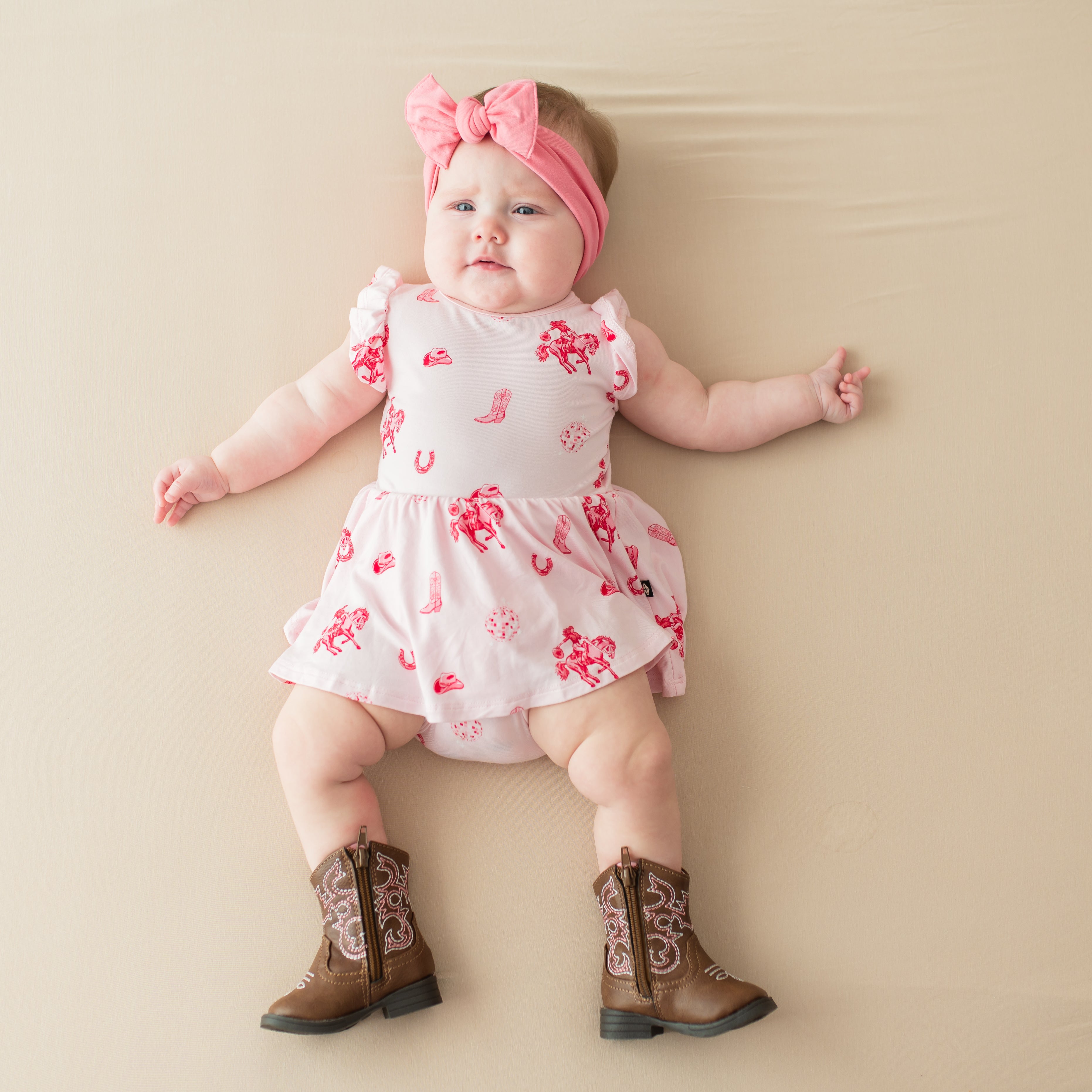 Infant laying on a light neutral background wearing the Twirl Bodysuit Dress in Disco Cowgirl with a knotted bow headband in guava and brown cowboy boots