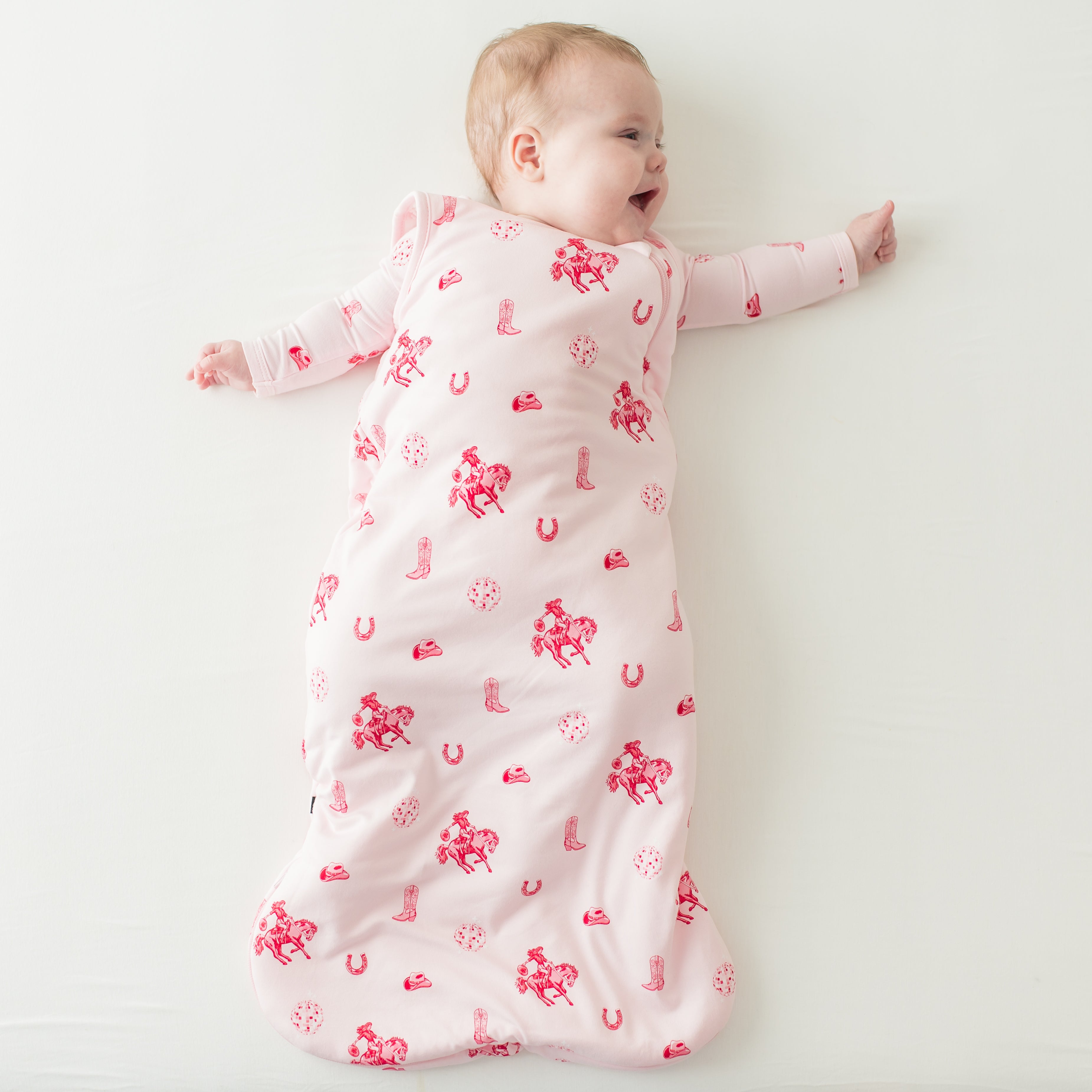Young infant girl laying on a cream surface wearing the Sleep Bag in Disco Cowgirl 1.0 and matching long sleeve romper