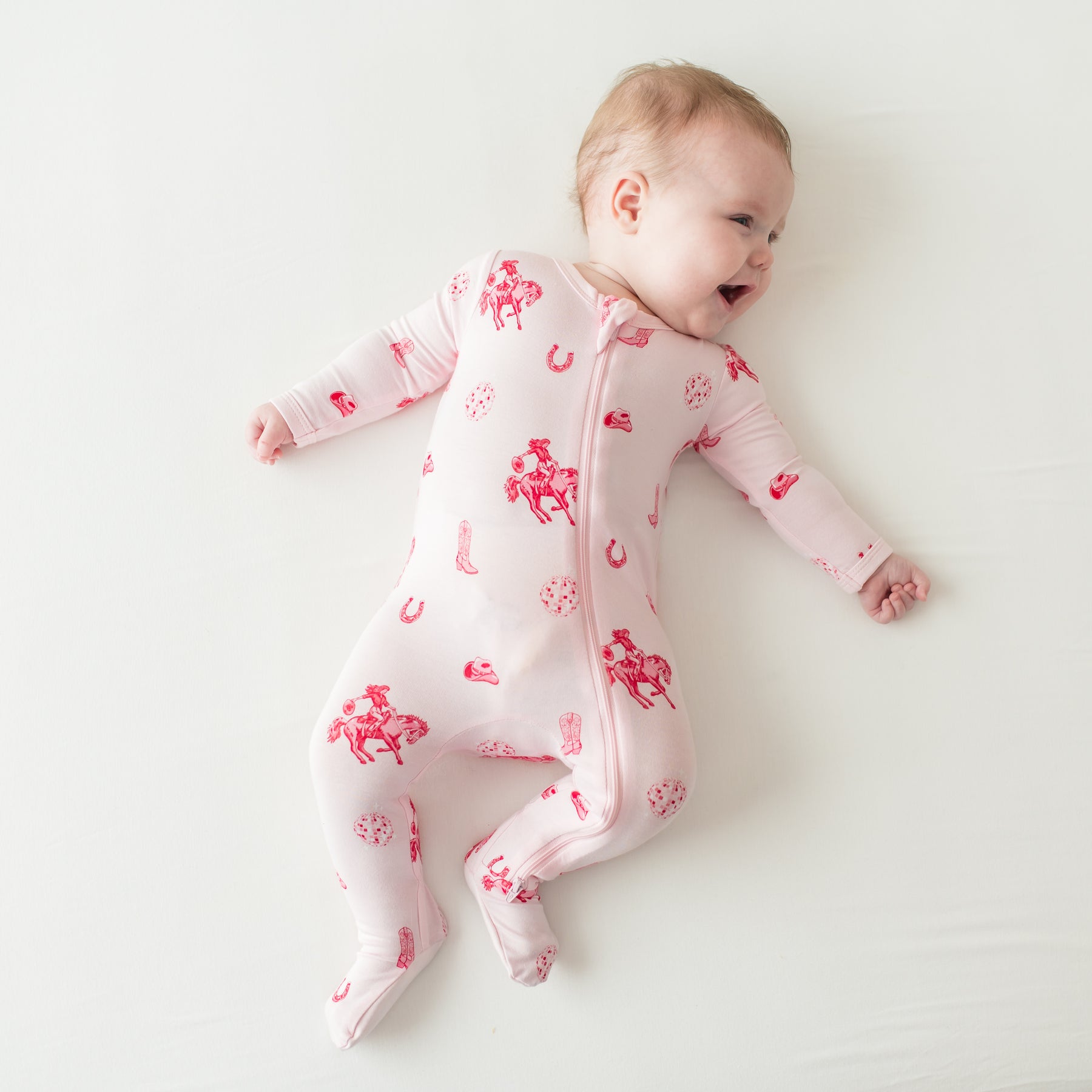 Young smiling infant laying on a cream surface wearing the Zippered Footie in Disco Cowgirl