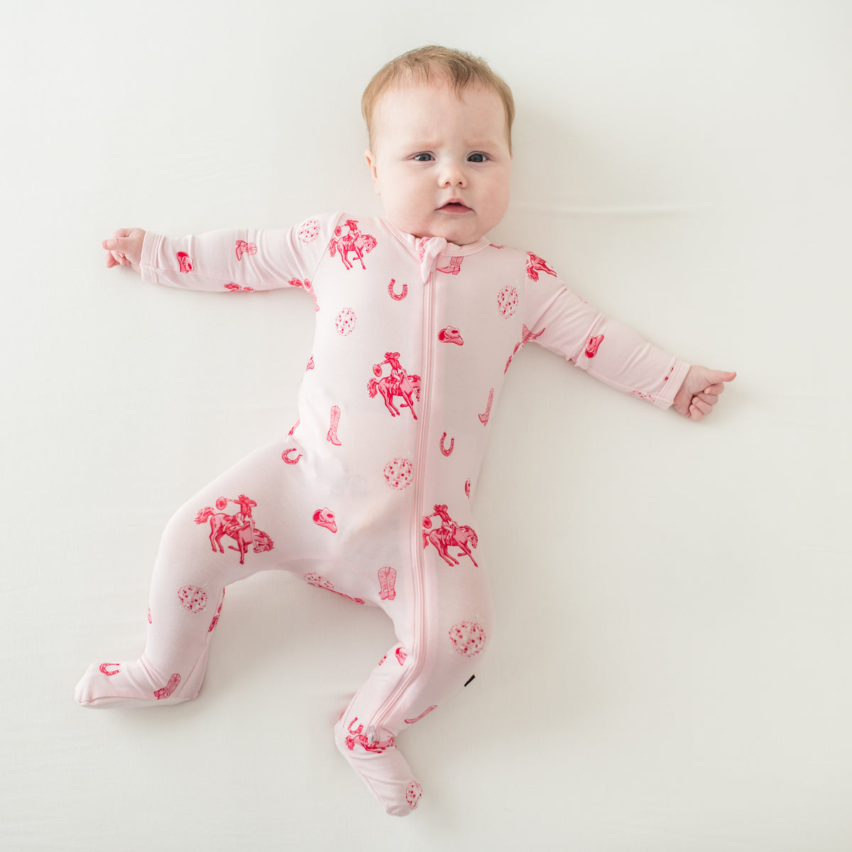 Infant girl laying on a cream surface stretched out wearing the Zippered Footie in Disco Cowgirl