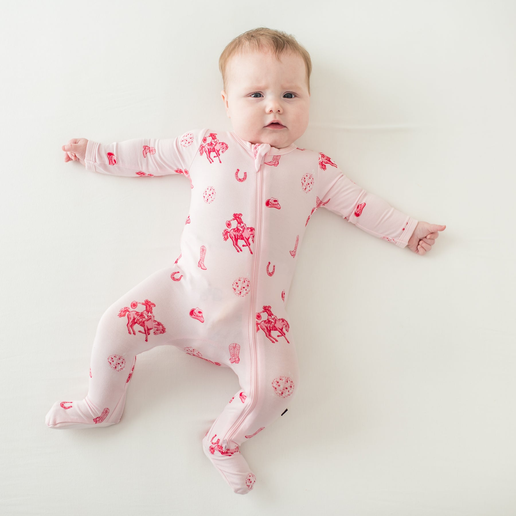 Infant girl laying on a cream surface stretched out wearing the Zippered Footie in Disco Cowgirl
