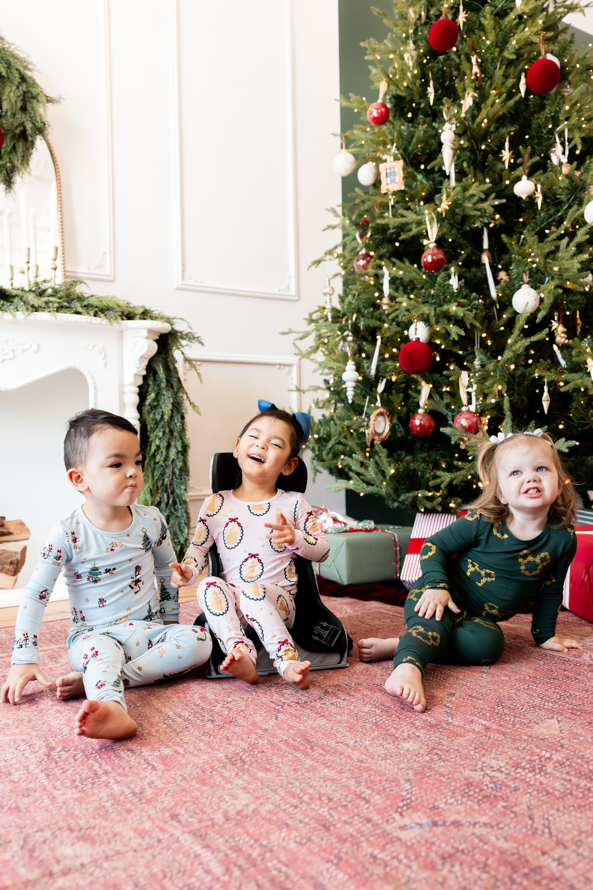 Three kids wearing Long Sleeve Pajamas in Mickey & Friends Christmas print, Mickey Christmas Wreath and Princess portrait sitting together smiling beside a Christmas tree